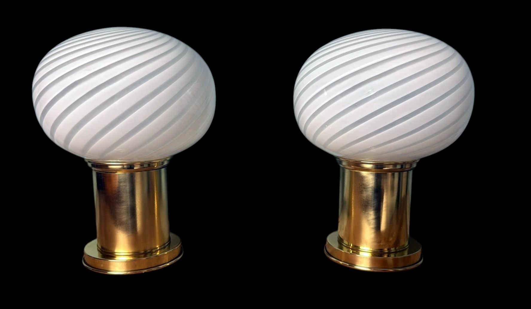 Elegante par de lámparas de sobremesa vintage con una esfera de cristal de Murano soplado a mano que descansa sobre una sólida base cilíndrica de latón. El cableado eléctrico discurre discretamente por la base, oculto bajo una cubierta metálica