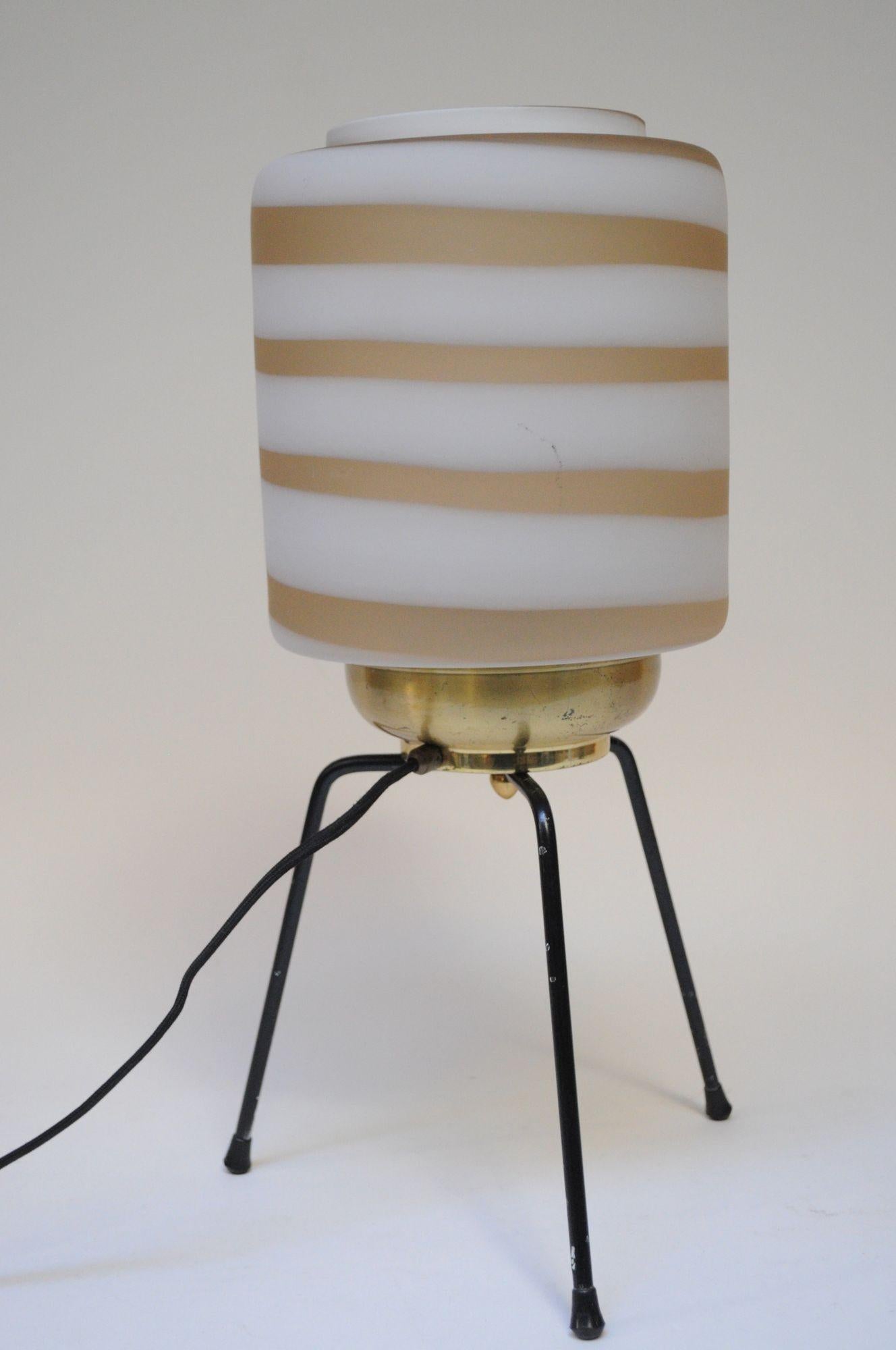 Impressive table lamp composed of a rubber/plastic tripod base supporting a brass cradle with Murano glass shade (ca. 1950s, Italy).
Frosted cylindrical glass shade is a swirl of taupe-beige against white and emits a warm, potent light.
Overall,