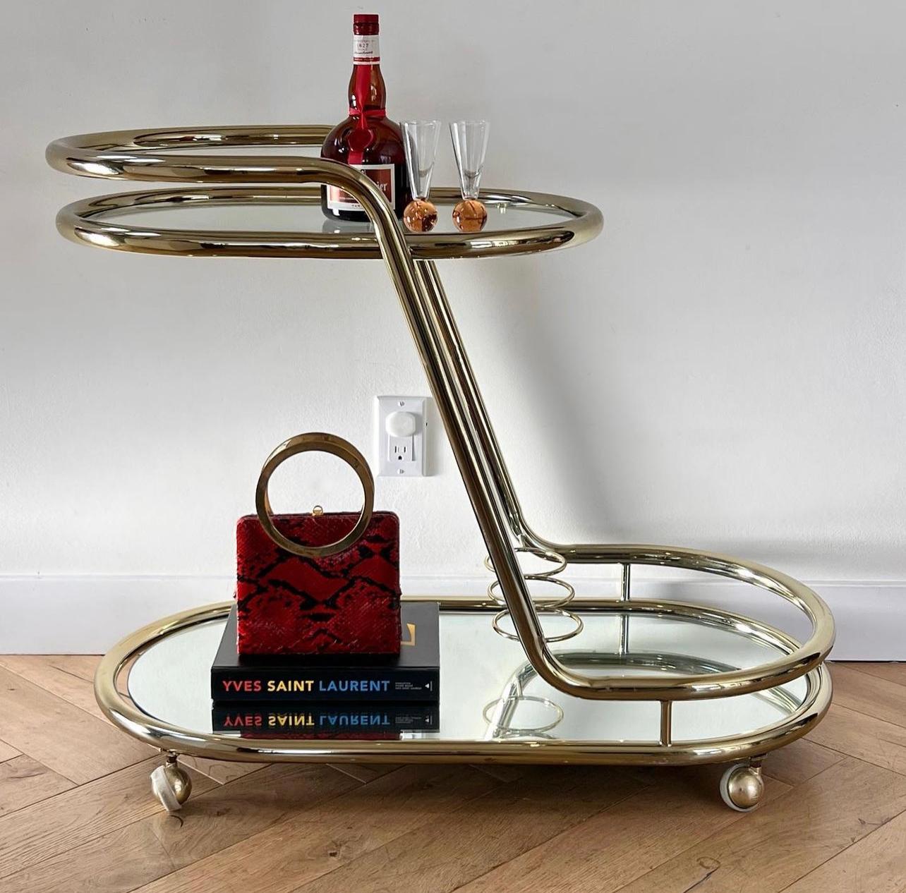 ITALICS: a midcentury two-tier tubular Italian bar cart attributed to Morex, circa 1970s. Featuring a slanted frame in subtly gold plated chrome with glass top and a mirror base, as well as bottle enclaves on the second tier. Both shelves are