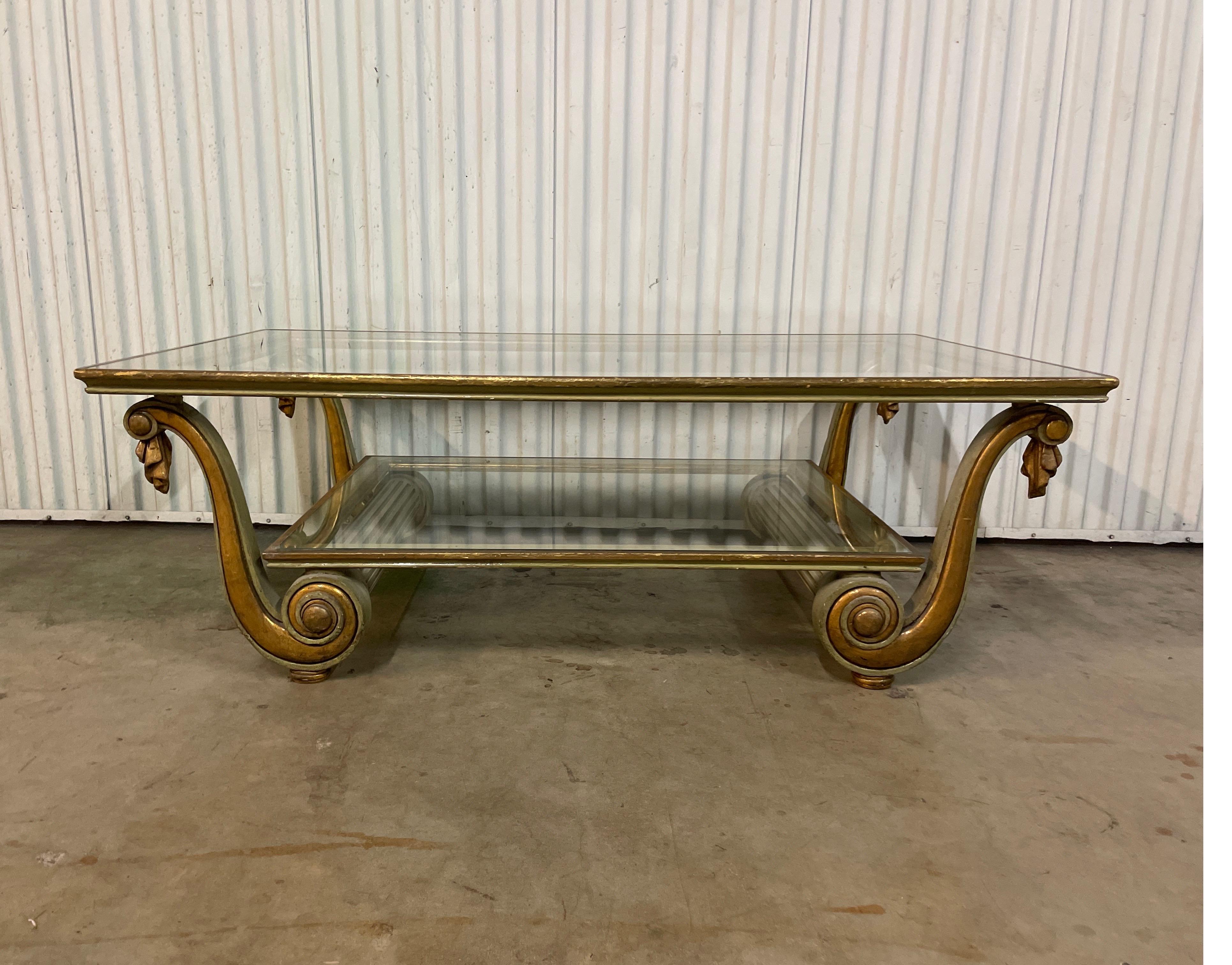 Hollywood Regency style cocktail table with two glass shelves. Frame is carved, painted & gilded. Sold as a single item but two are available.