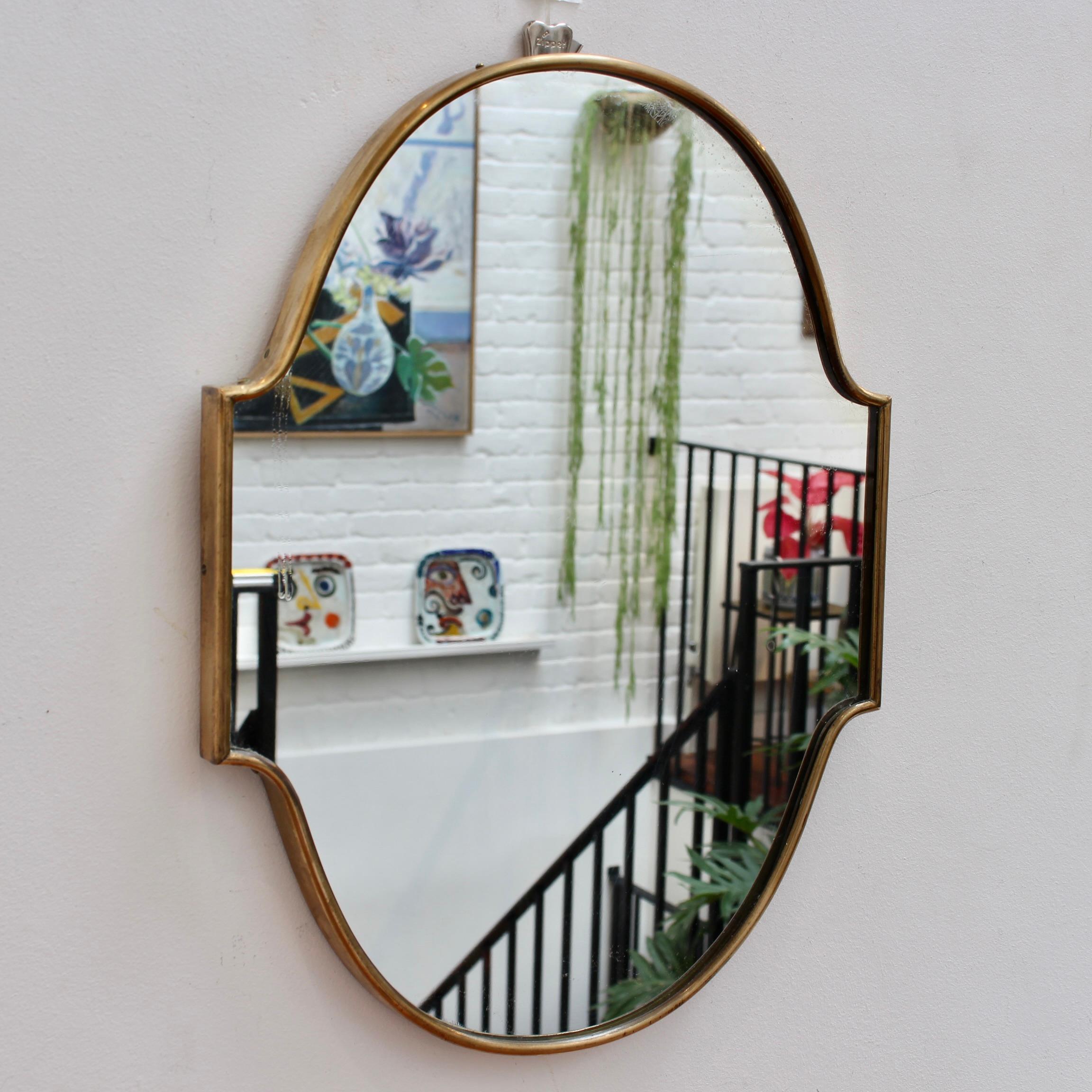 Small-scale midcentury Italian wall mirror with brass frame (circa 1950s). The mirror is oval-shaped with 'ears' on each side. It is classically elegant and distinctive in a modern Gio Ponti style. The mirror is in overall good condition with some