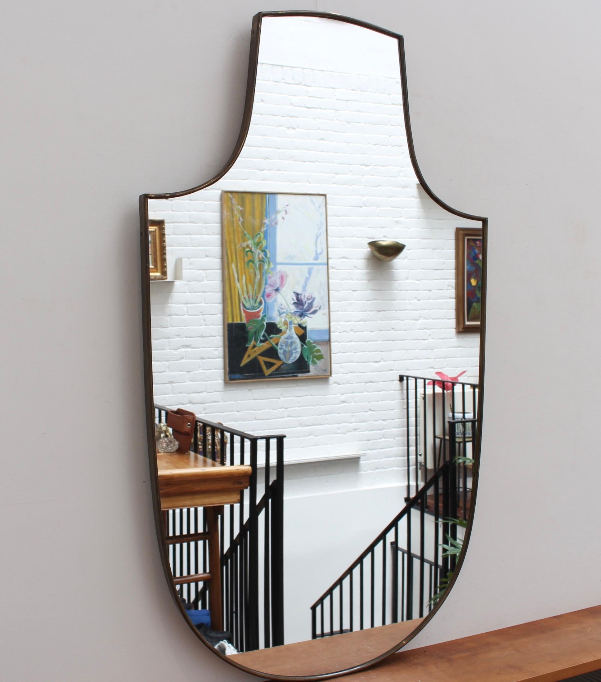 A large Mid-Century Italian wall mirror with brass frame (circa 1960s). The mirror is classically-shaped and distinctive in a Modern style. It is in fair overall condition. A beautiful age-related patina develops on the brass frame along with some