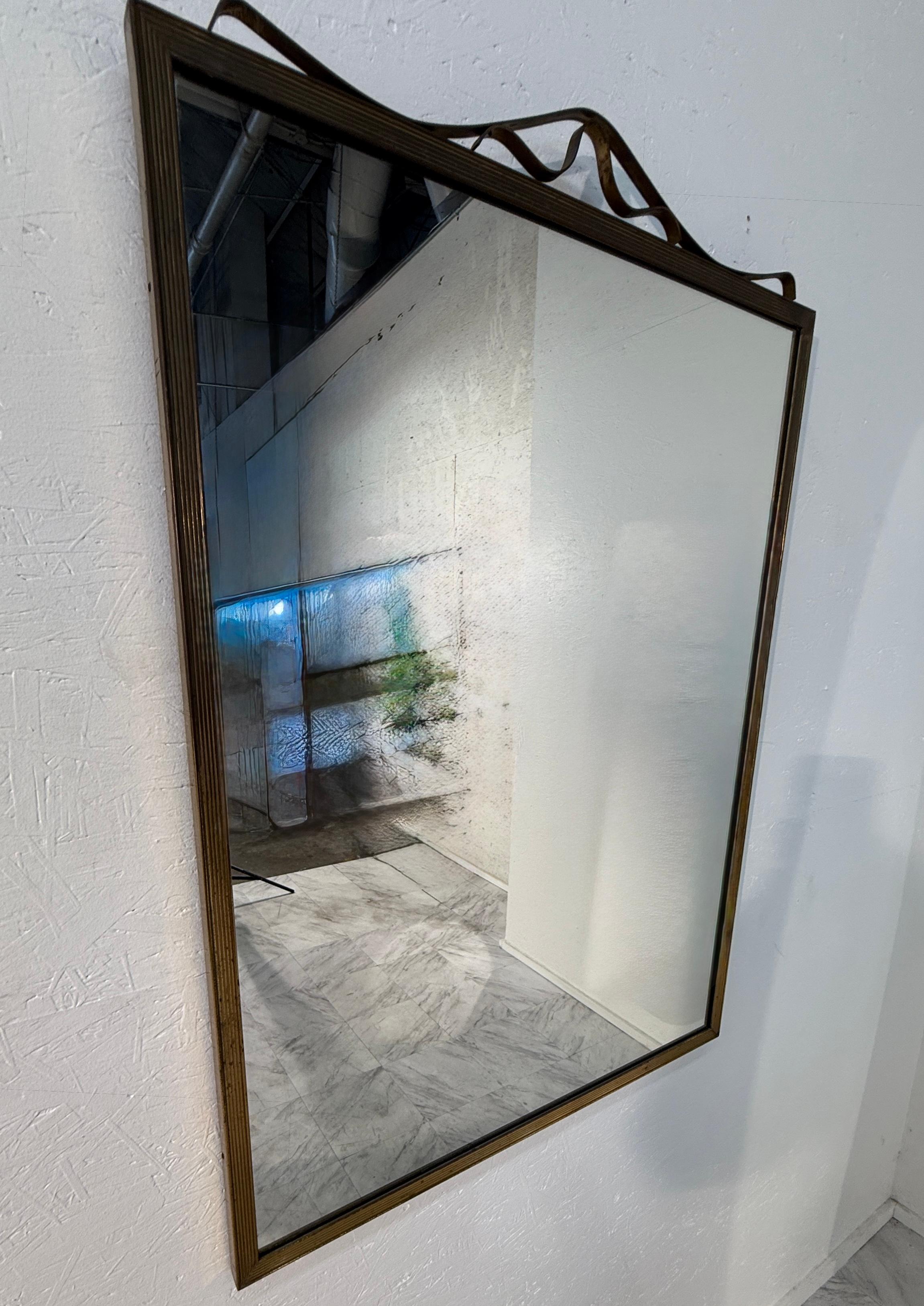 Specchio rettangolare da parete vintage con cornice in ottone finemente rigata e un dettaglio decorativo a nastro metallico ondulato lungo il bordo superiore. La cornice presenta una patina naturale e un invecchiamento che contribuisce al suo