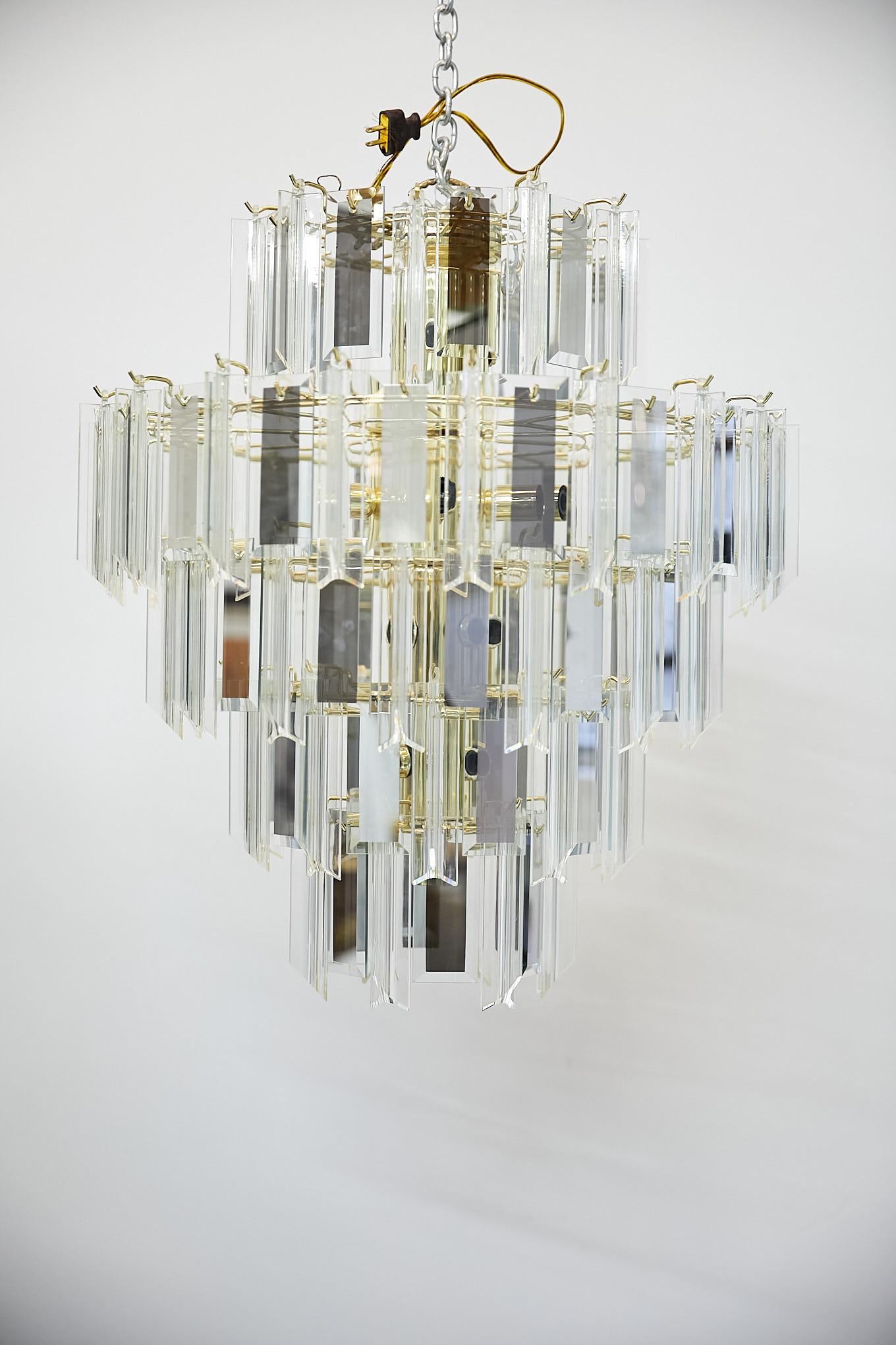 Vintage Italian five-tier waterfall or wedding cake style chandelier having a cylindrical brass frame and fittings.
The chandelier has alternating clear Lucite prisms and rectangular mirrored and beveled glass panes that cascade and taper downward.