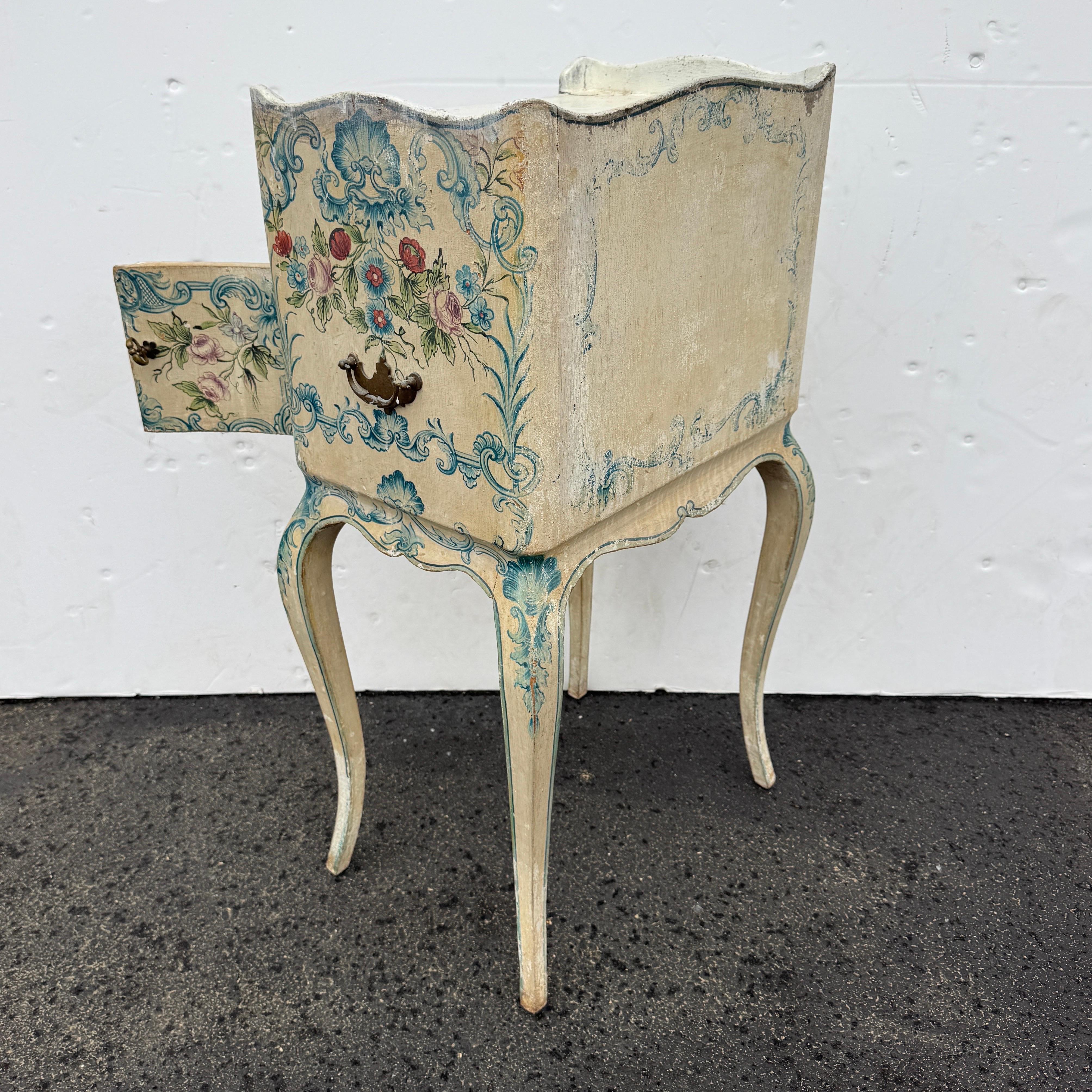 Fabulosa mesilla de noche con motivos florales pintados en cremas, rojos y azules. Esta mesa es una pieza muy versátil que puede utilizarse como mesa auxiliar en un salón, recibidor o junto a una cama en cualquier ambiente doméstico formal o