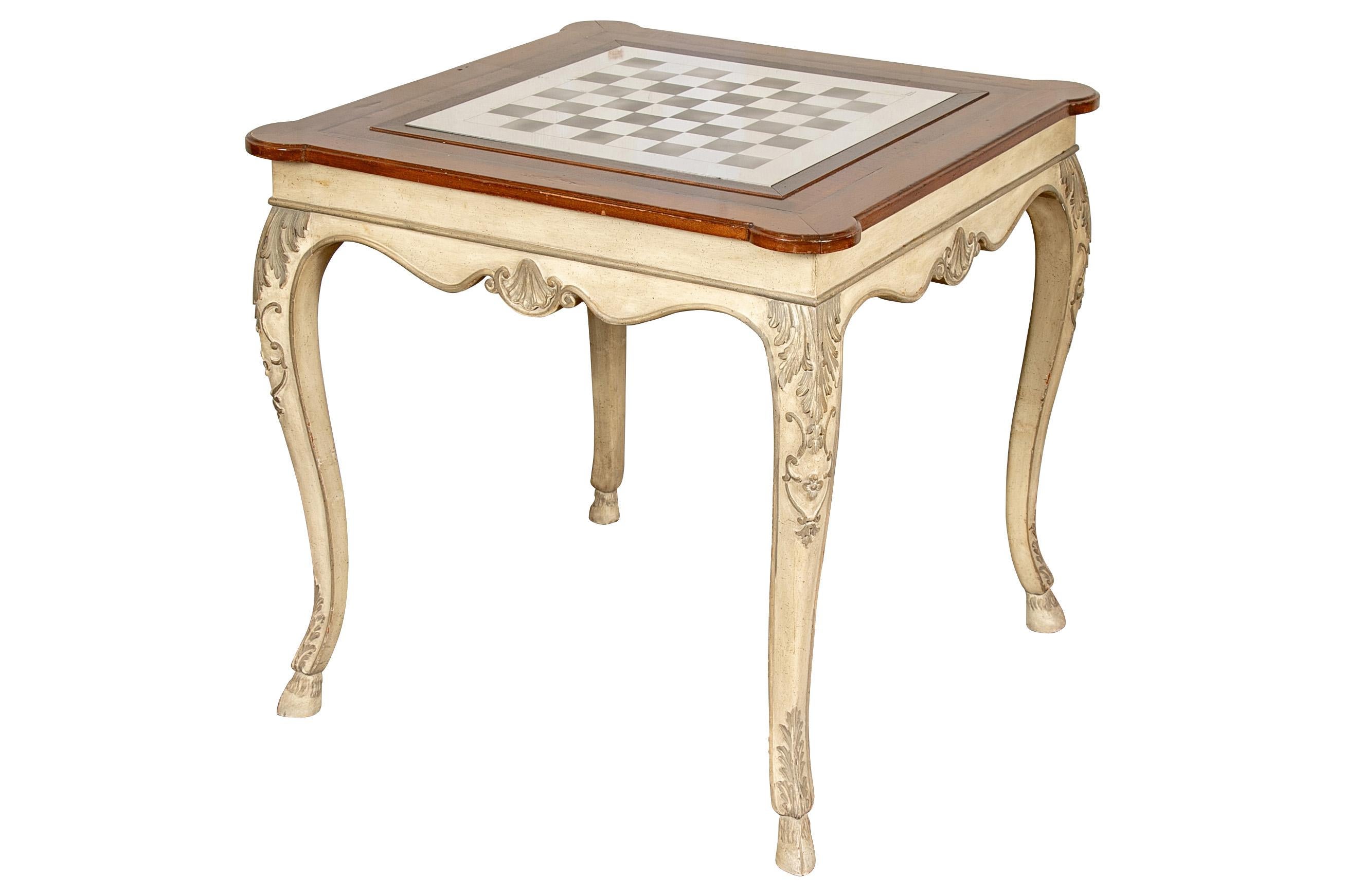 A banded table with cookie corners loose on the carved frame painted in sage green on cream with leafy details and raised on hoof feet. The top with inset shiny and brushed chrome chess board signed Cenedese Murano, along with black and clear glass