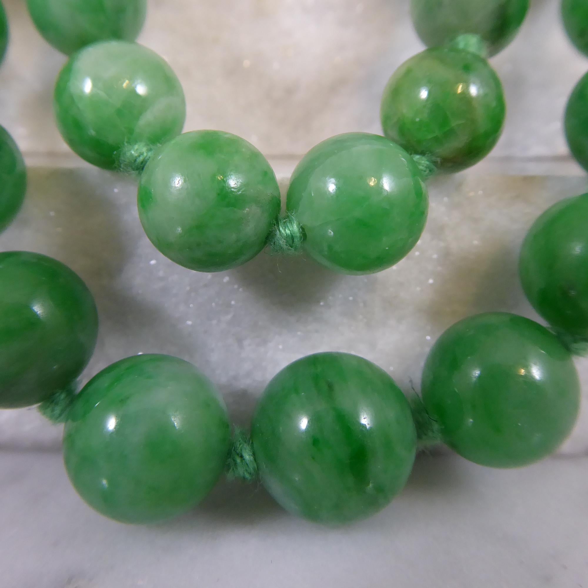 A jade bead necklace comprising two rows of graduating sized, spherical green jadeite beads measuring between 4.15mm and 9.80mm diameter.  One hundred and twelve beads in total strung knotted to green cotton string and attaching to a double rounded,