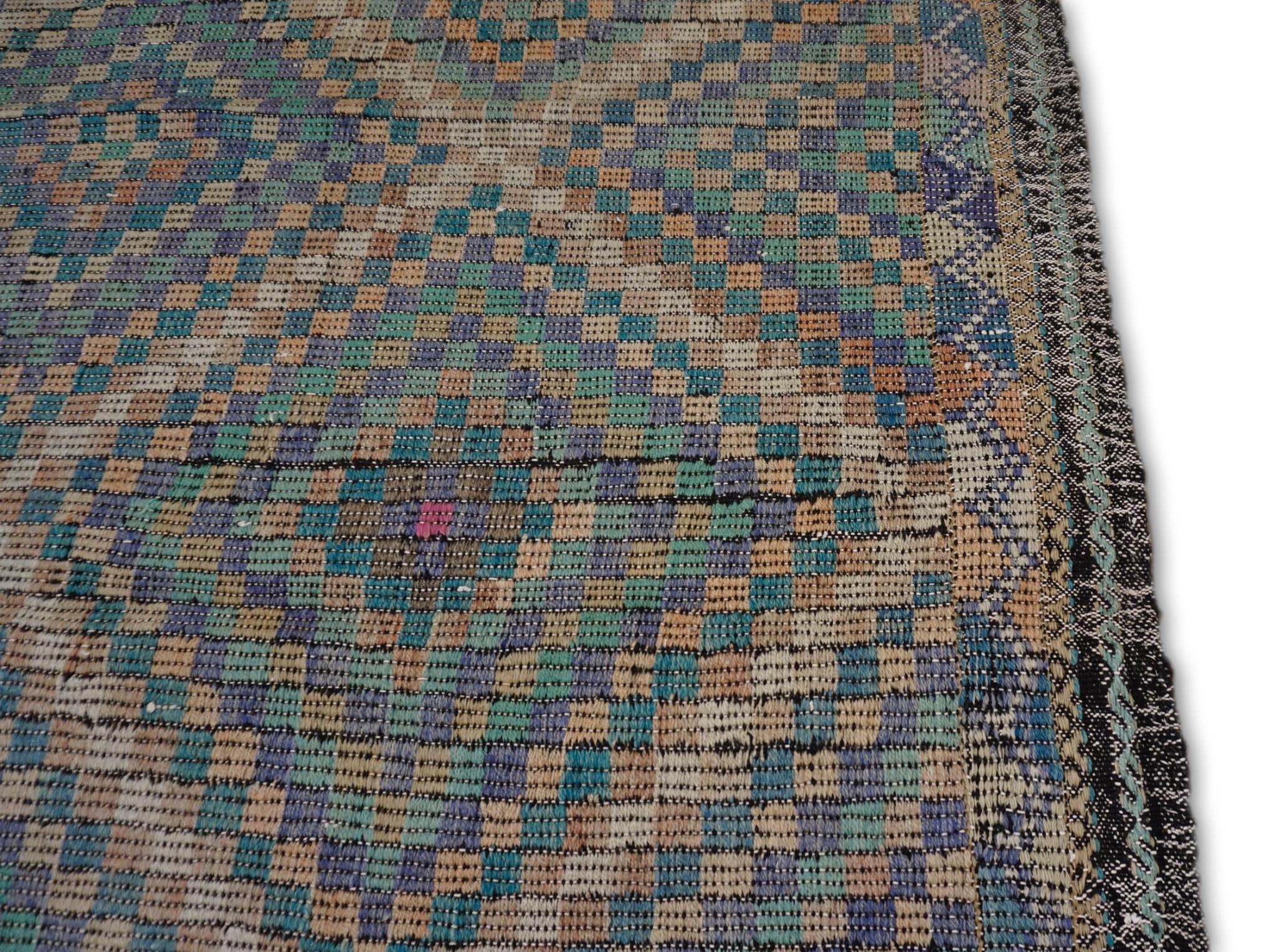 This vintage wool kilim is a captivating blend of traditional craftsmanship and timeless design. Showcasing an intricate geometric diamond pattern across its surface, the rug is adorned with green, gray, tan, and purple. The rhythmic arrangement of