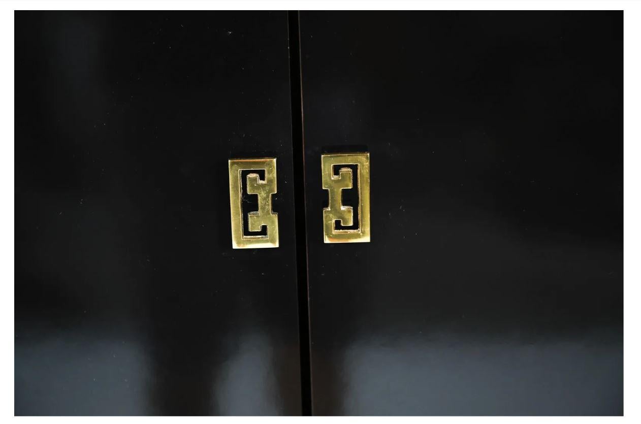 A vintage black lacquer James Mont style cabinet with Asian motif brass hardware. Two large top doors open to reveal interior cubbies and three drawers open below, each with double Asian motif pulls. Cabinet sits on a brass base.