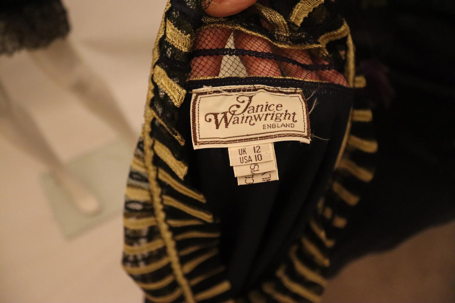 Un vestido de noche vintage de la diseñadora británica Janice Wainwright. El vestido es de un tejido negro y dorado con rayas negras y doradas en el cuerpo y detalles cruzados negros y dorados en las mangas. El cuerpo del vestido está forrado y  Hay