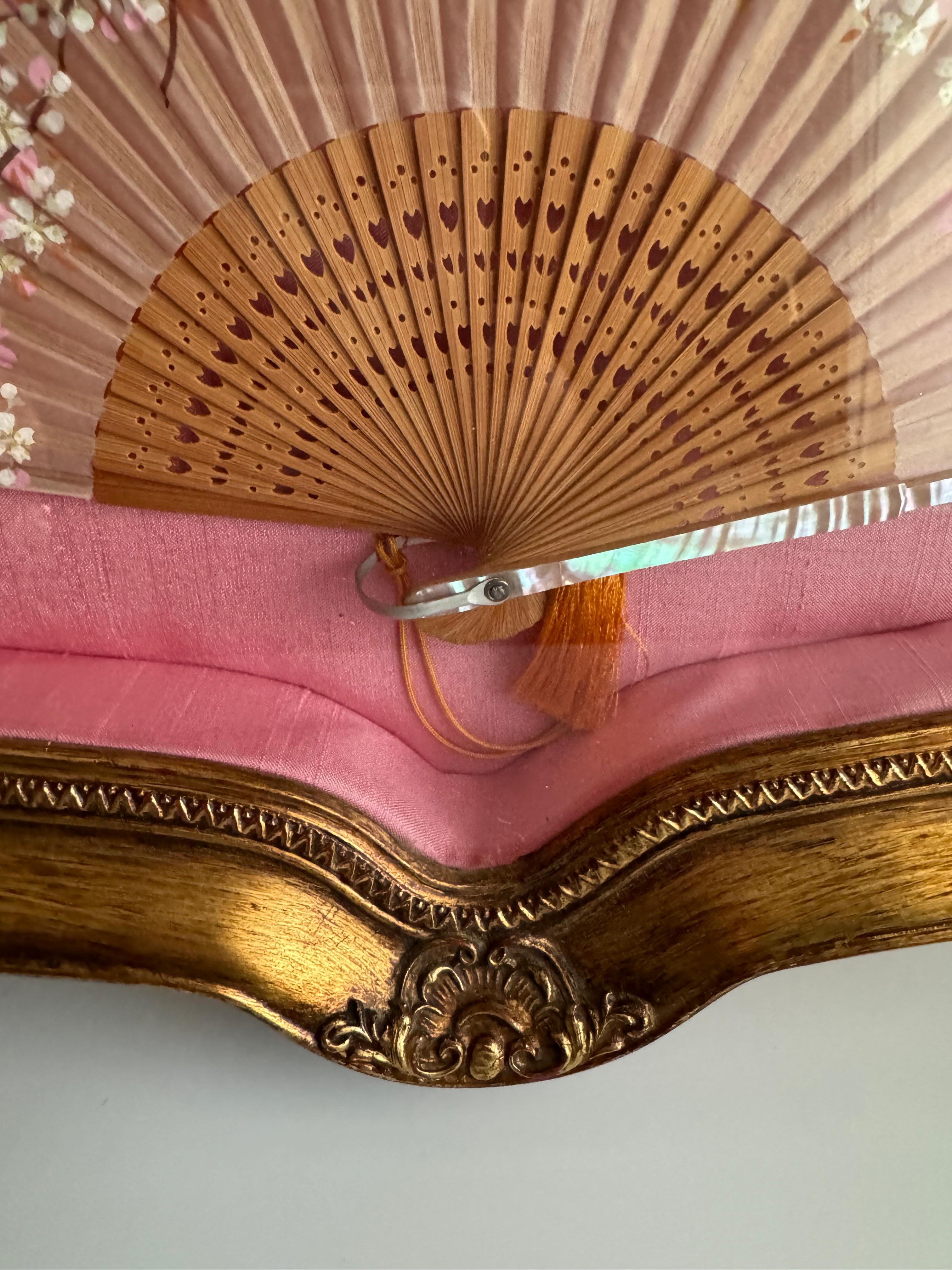Gorgeous Japanese vintage fan . It features hand-painted Cherry Blossom details and inlaid mother of pearl elements. The fan is displayed within a shadow box frame, which seems to be lined with pink silk, enhancing its aesthetic appeal. The frame