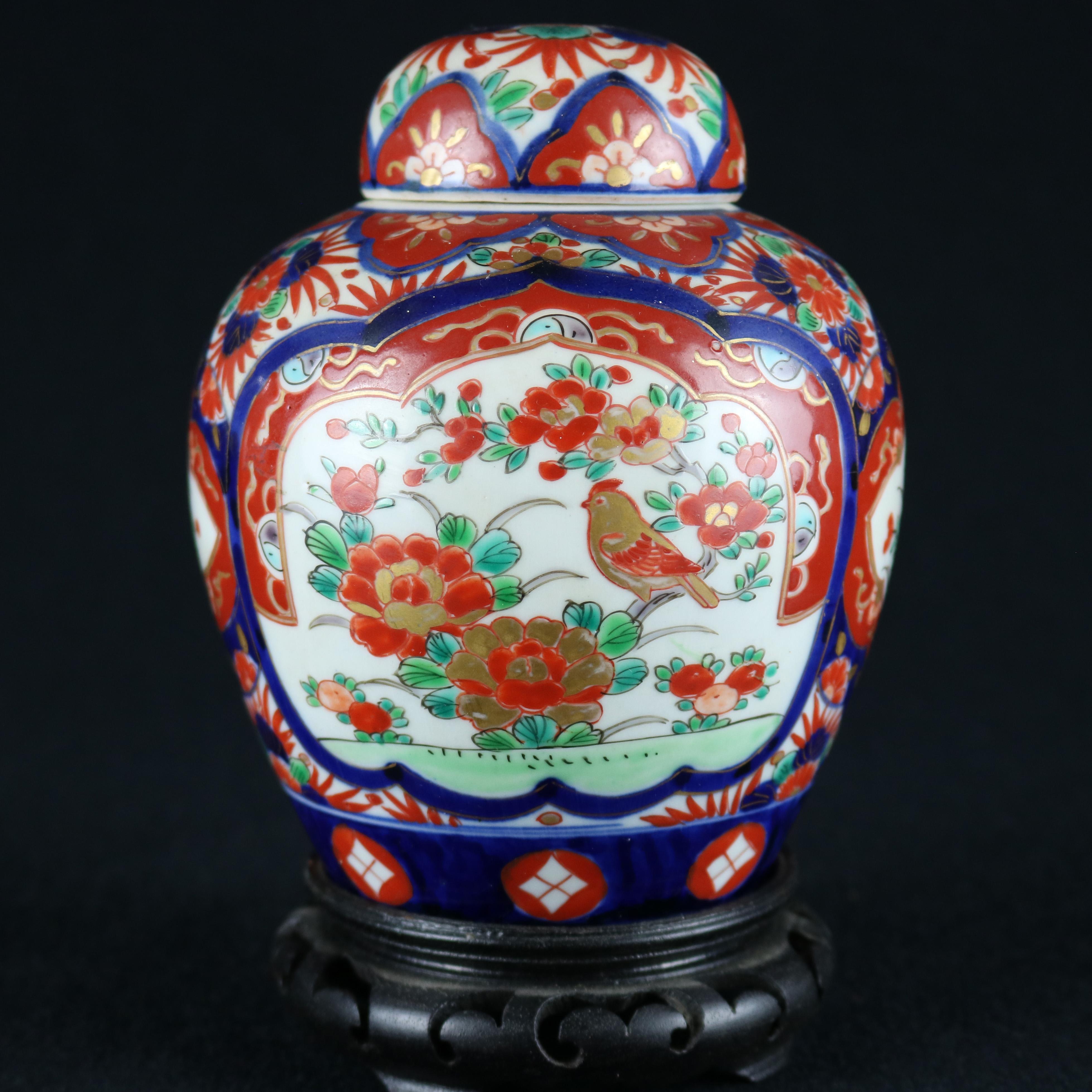 A vintage Japanese Imari covered ginger jar offers porcelain construction with hand enameled decoration including reserves with floral elements, seated on ebonized carved hardwood base, stamped Made in Japan on base, 20th century.

Measures: 7.25