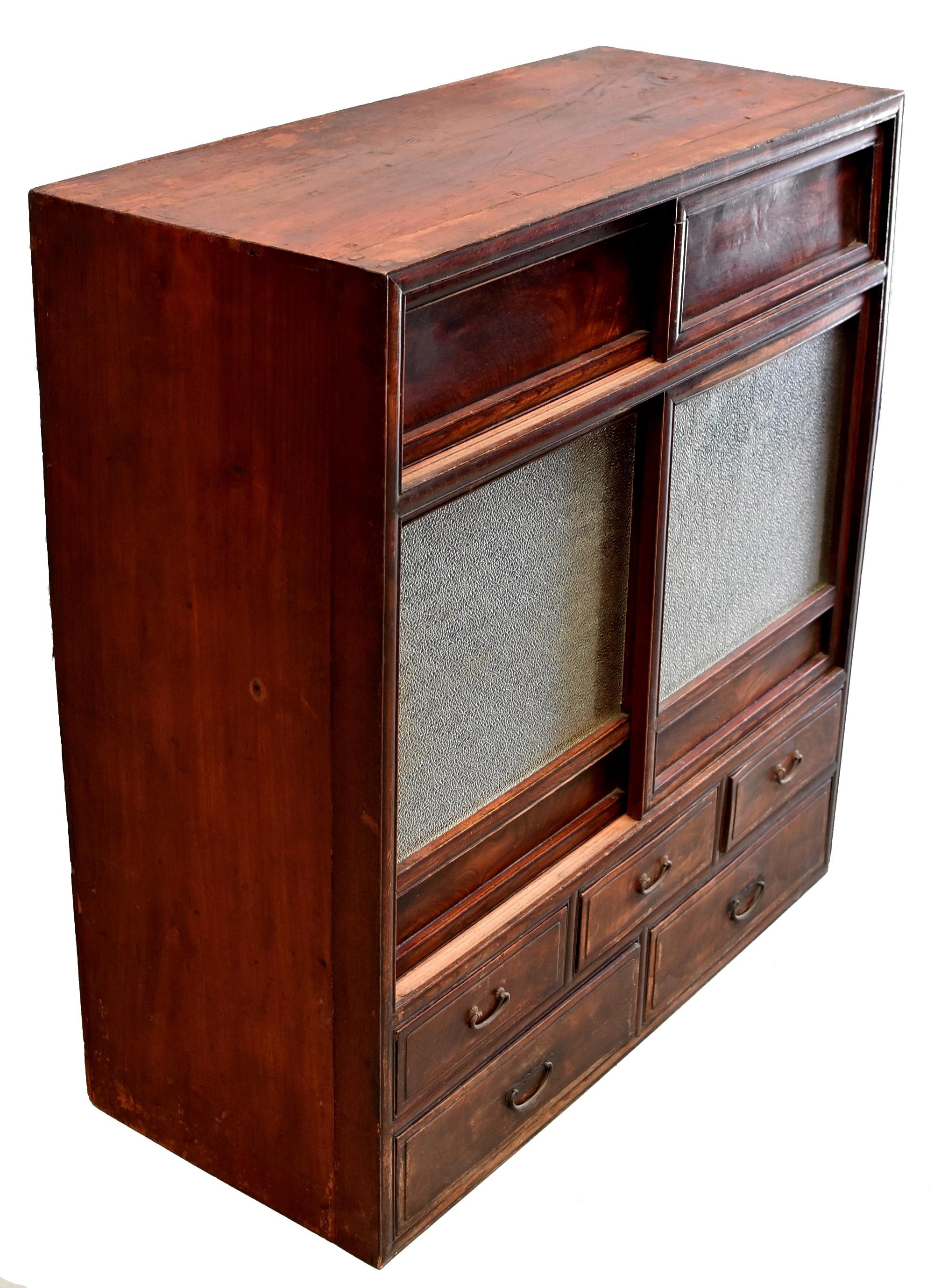 A beautiful vintage Japanese Tansu with five drawers and a pair of each glass and wood sliding doors. Behind the textured glass doors, is a tiered shelf unit with a nice carved feature. The wood has beautiful grains. Five full capacity drawers offer