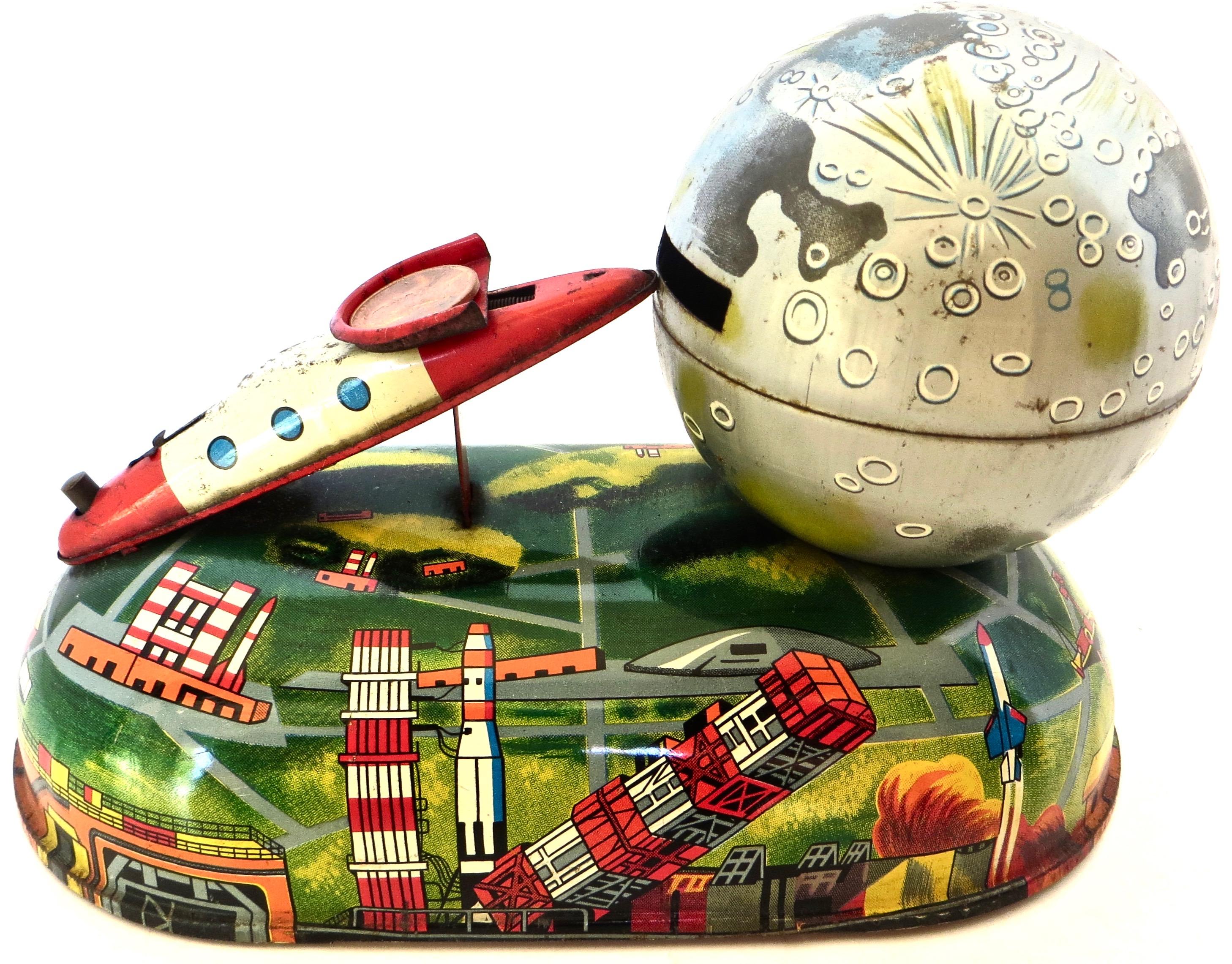 Japanese tin mechanical bank features a rocket launcher mounted on the top surface of the body which fires a missile (penny or coin) into outer space, as represented by a globe like sphere. The graphics to the base and sphere are quite bright,