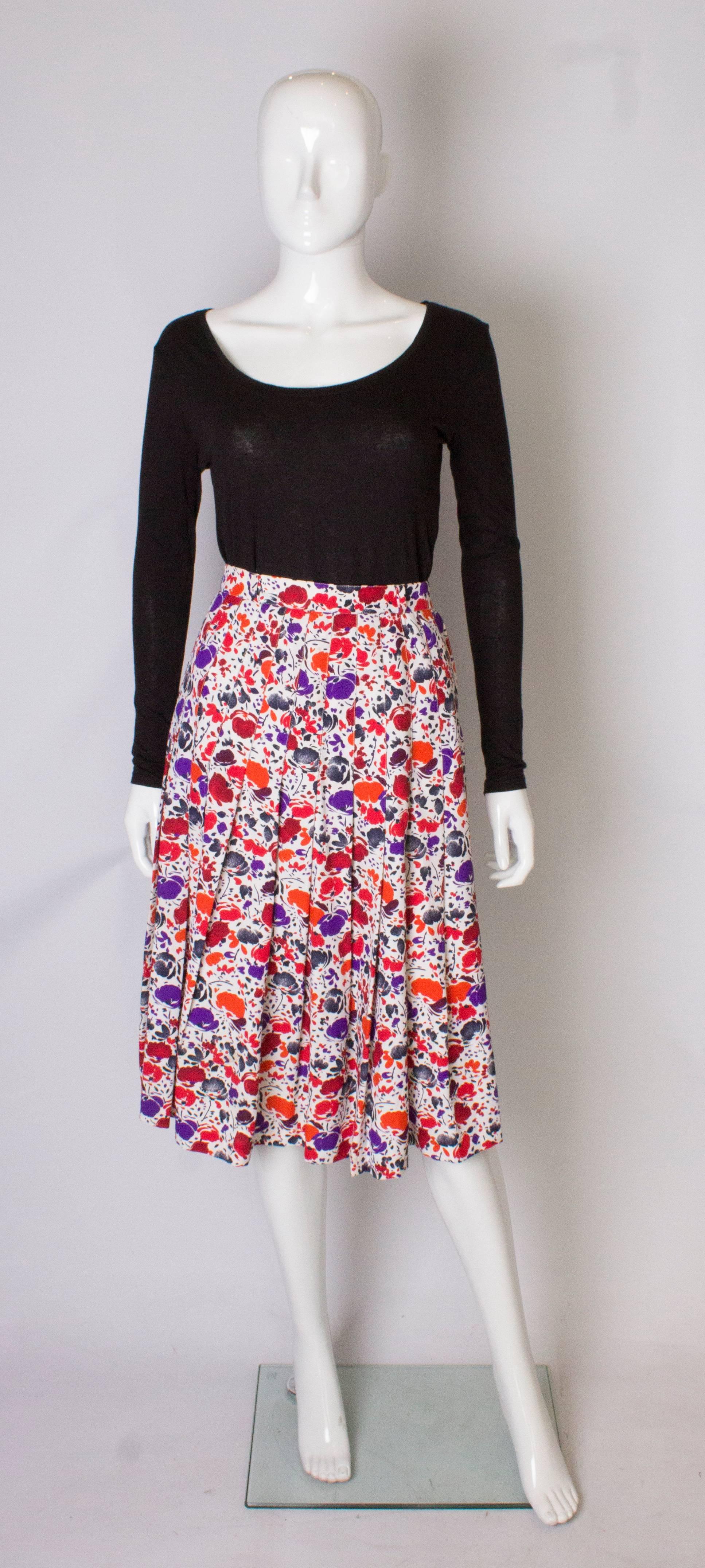 A chic vintage pleated skirt by Jean Louis Scherer Boutique. The skirt is number 087951, and has a white background with a red, orange and purple print. The skirt has sewn in pleats to the hip and so creates a smooth line. The skirt has a zip