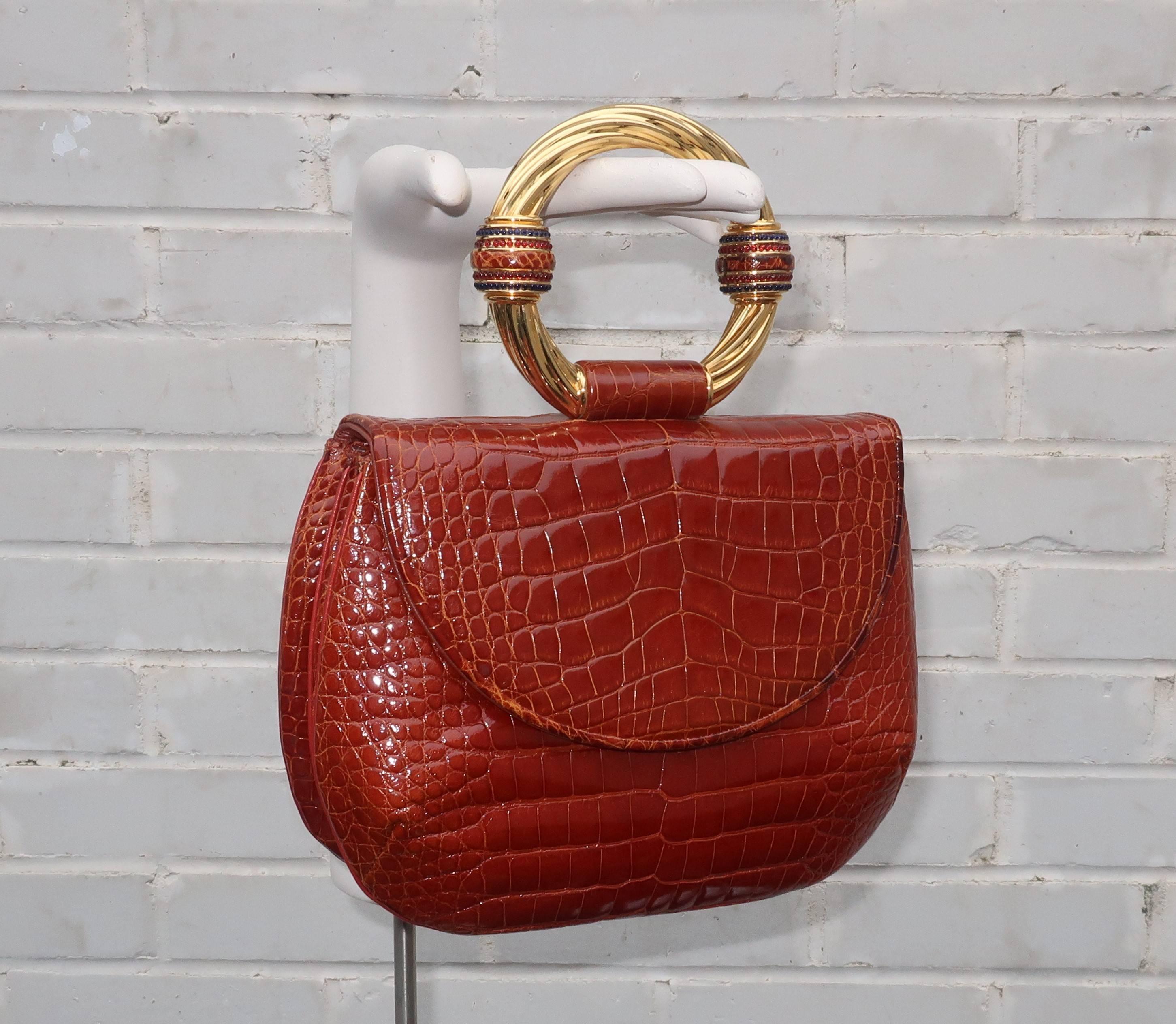 This stunning Judith Leiber alligator handbag serves double duty as a piece of costume jewelry for the wrist.  The versatile cognac colored skin is embellished with a large ring shaped gold tone metal handle.  The handle is decorated with skin and