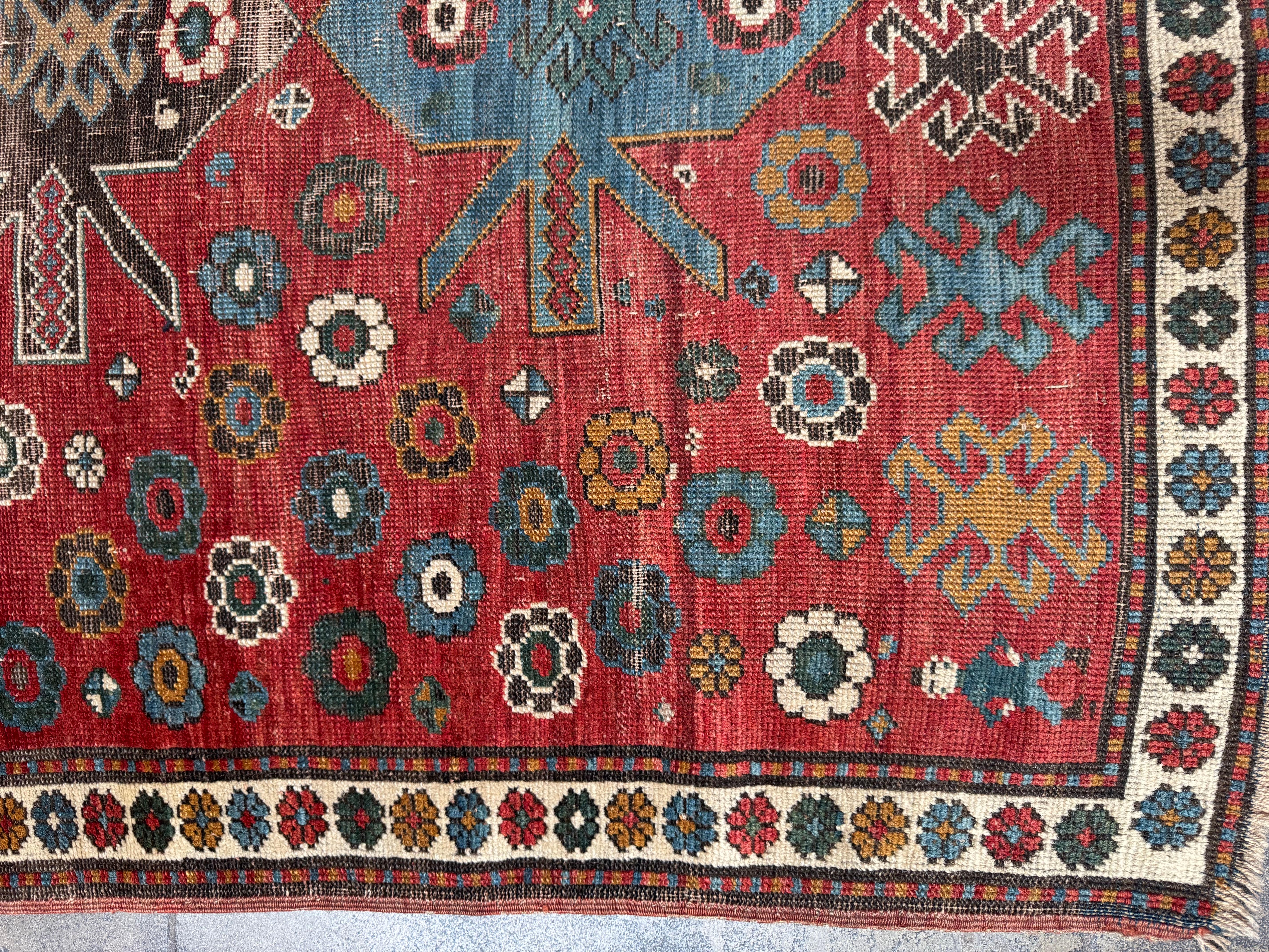 This vibrant vintage Kazak rug features a unique composition of floral rosettes and tribal medallions, set against a deep red ground that brings warmth and character to any space. The geometric star and diamond motifs in soft blue, ivory, camel, and