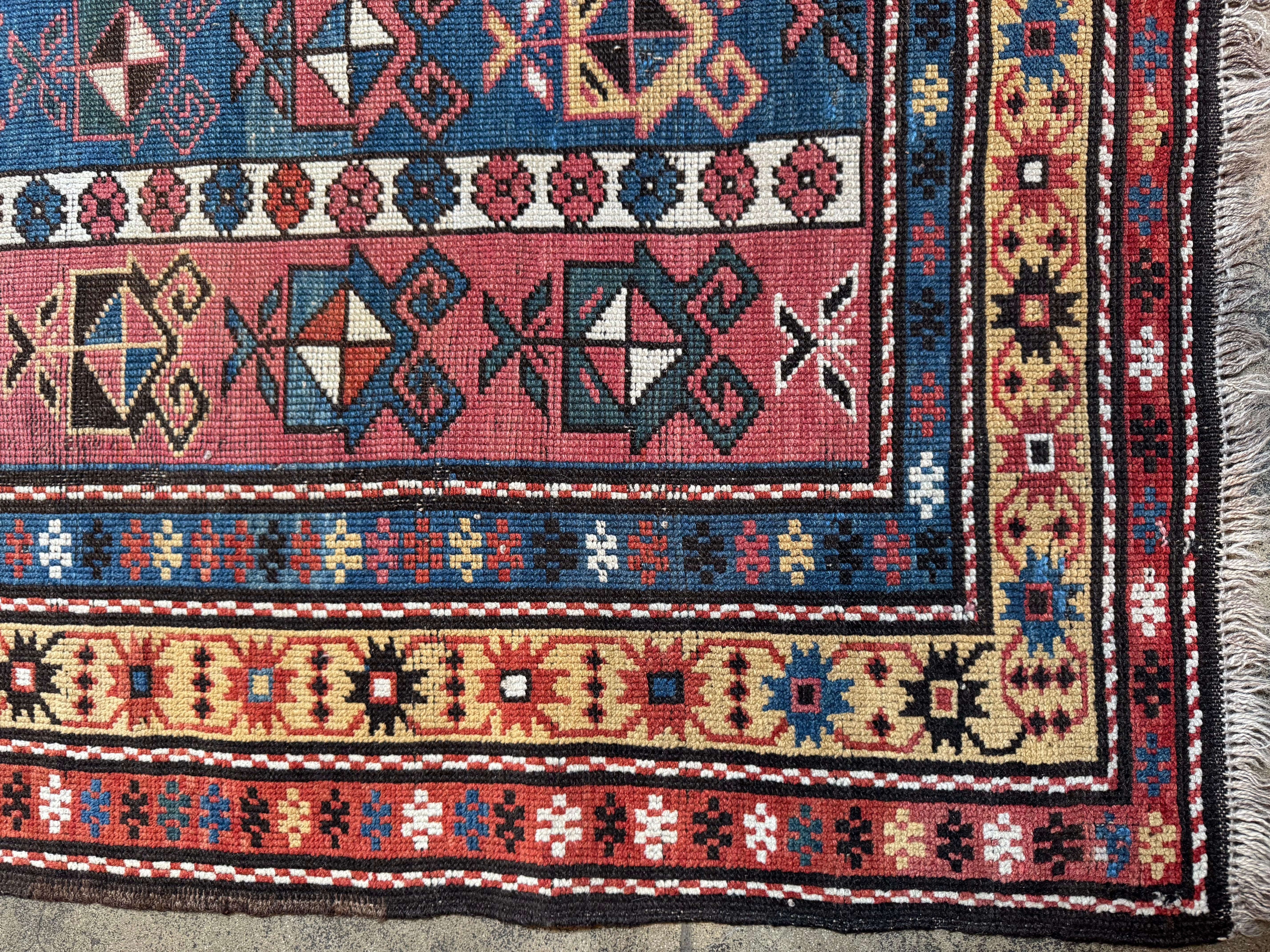 This vintage Kazak rug features a vibrant display of tribal artistry, showcasing bold geometric motifs arranged in structured columns over a deep red and indigo field. Flanked by multicolored borders rich in symbolic shapes and traditional Armenian
