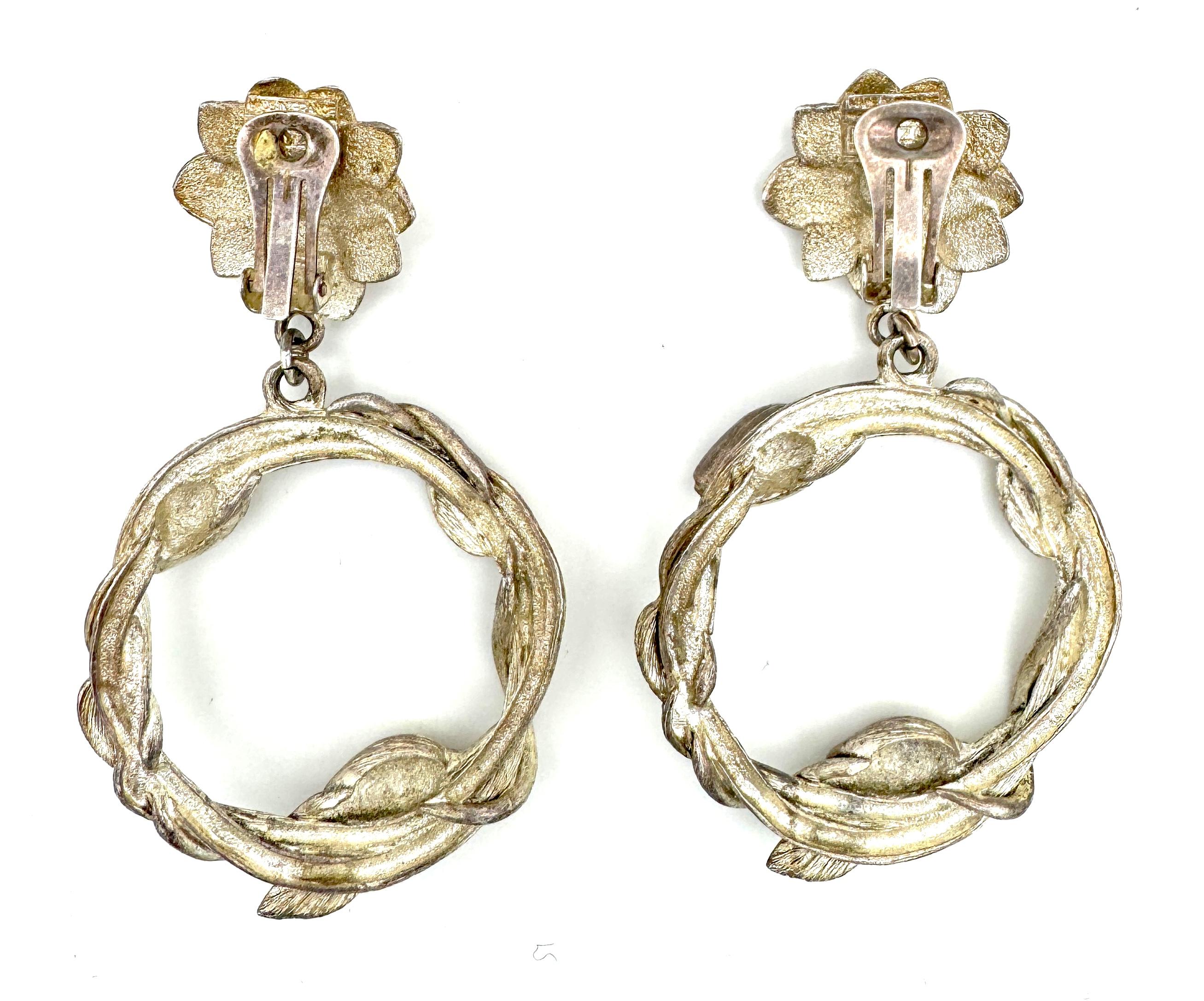 Estos sustanciosos pendientes de clip vintage fueron creados por Kenzo de París en los años 80 a partir de metal.
La parte superior de los pendientes está diseñada como flores de las que cuelga una corona hecha con una enredadera con capullos de