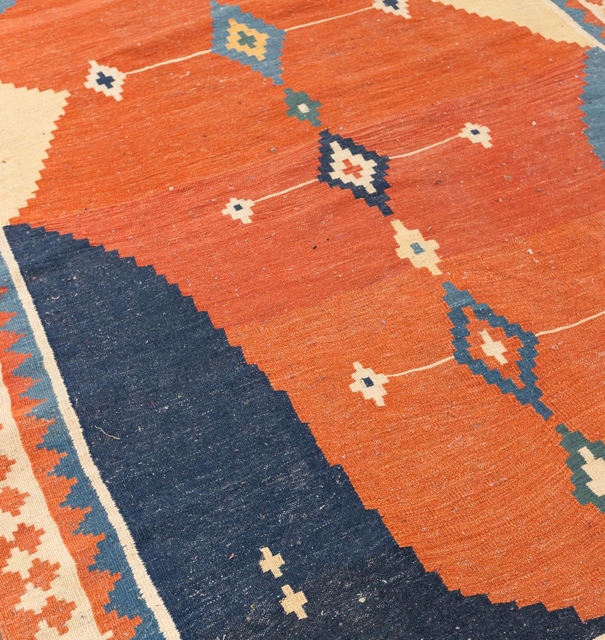 This vintage kilim offers a striking study in colour and geometry, defined by a bold terracotta field intersected by a sequence of diamond medallions in indigo, ivory and soft green. Its flatweave construction gives the surface a refined