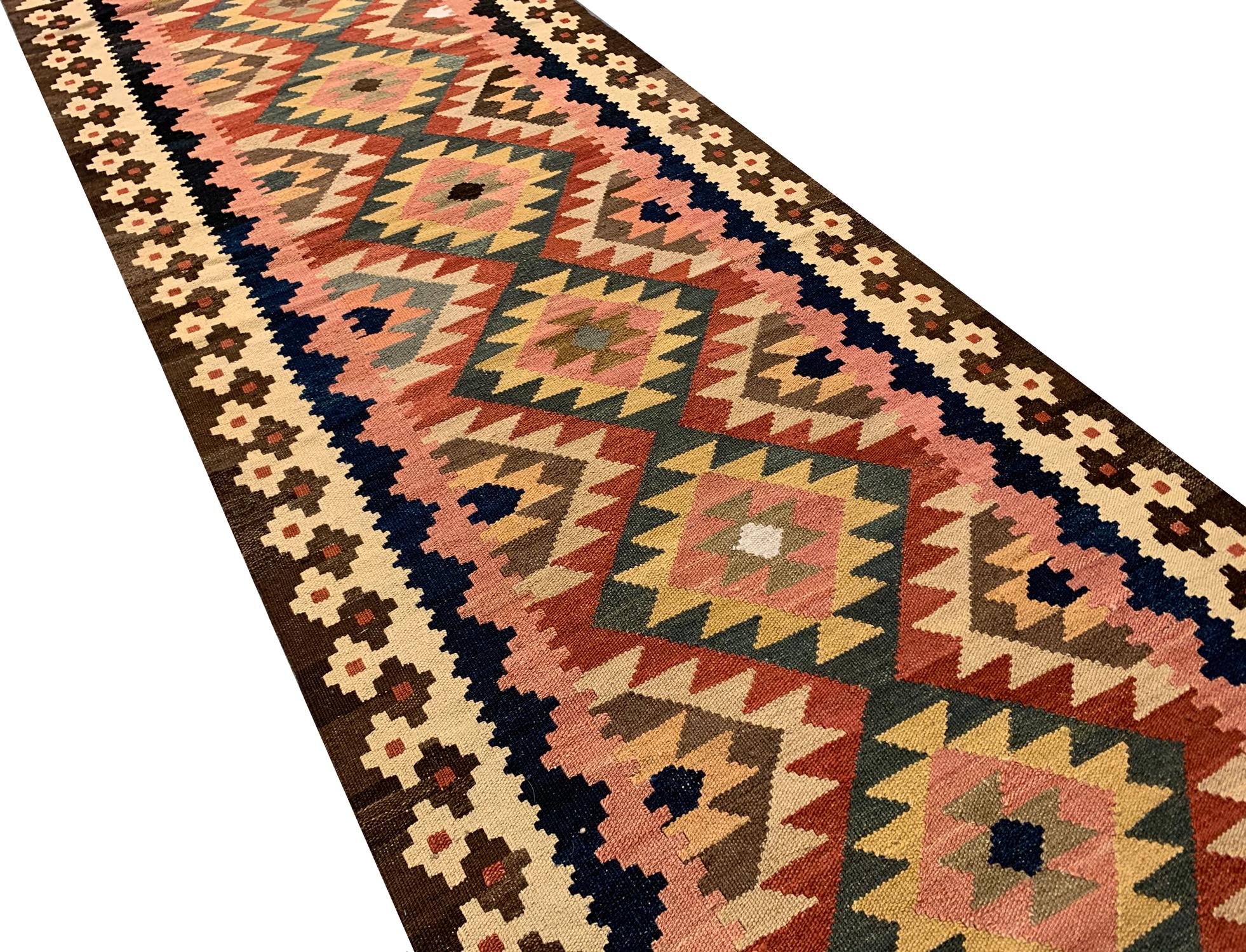 Ce Kilim Runner en laine fine est un tapis Kilim afghan moderne à tissage plat construit au début des années 2000. Le dessin présente un motif géométrique répété, tissé avec une palette de couleurs vibrantes, dont le vert, le rouille, le beige et le