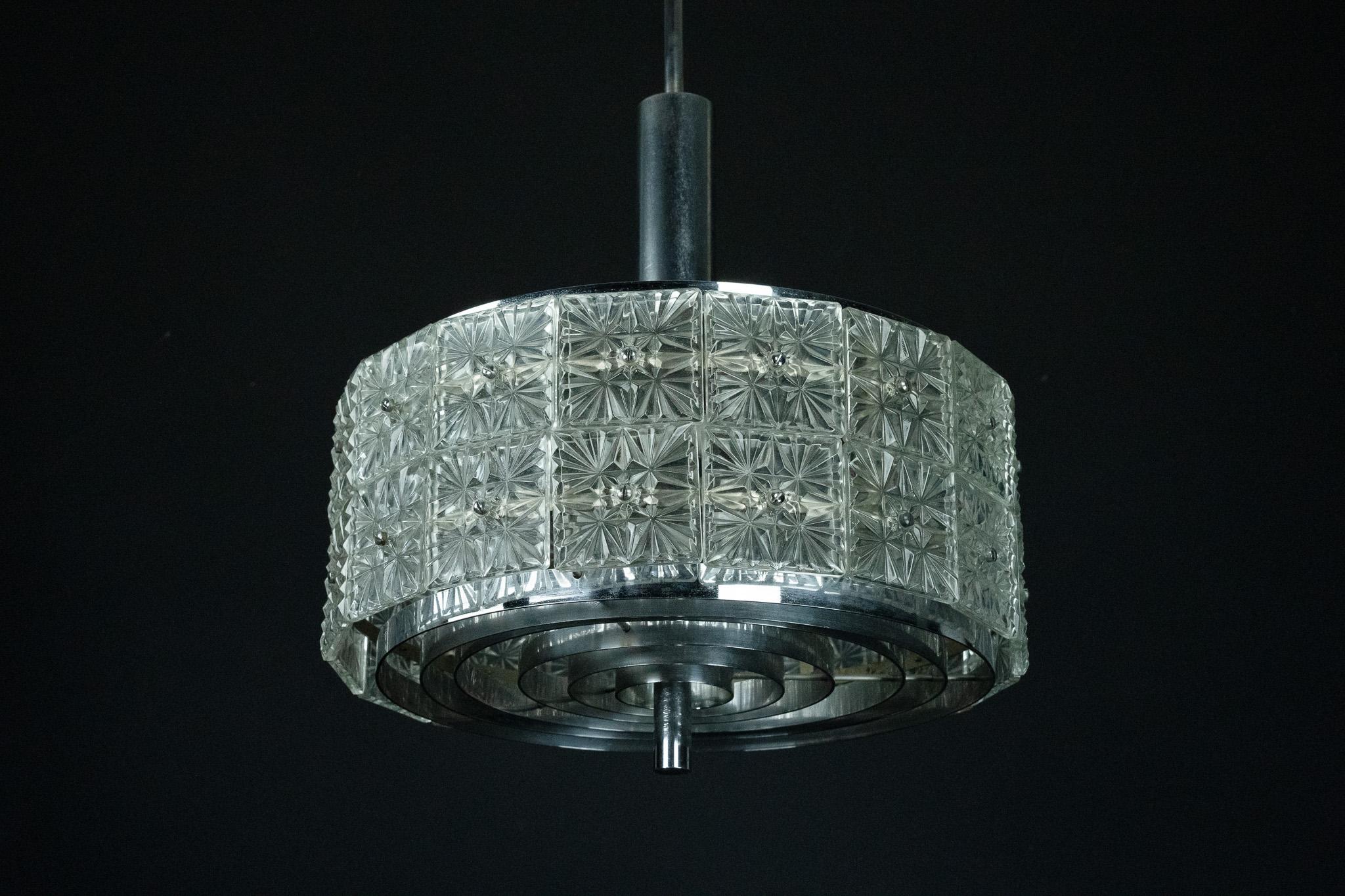Questo splendido lampadario da soffitto è probabilmente una produzione Kinkeldey degli anni '60, con una struttura geometrica realizzata in metallo cromato e pannelli in cristallo sfaccettato di alta qualità. Il pezzo combina l'eleganza moderna