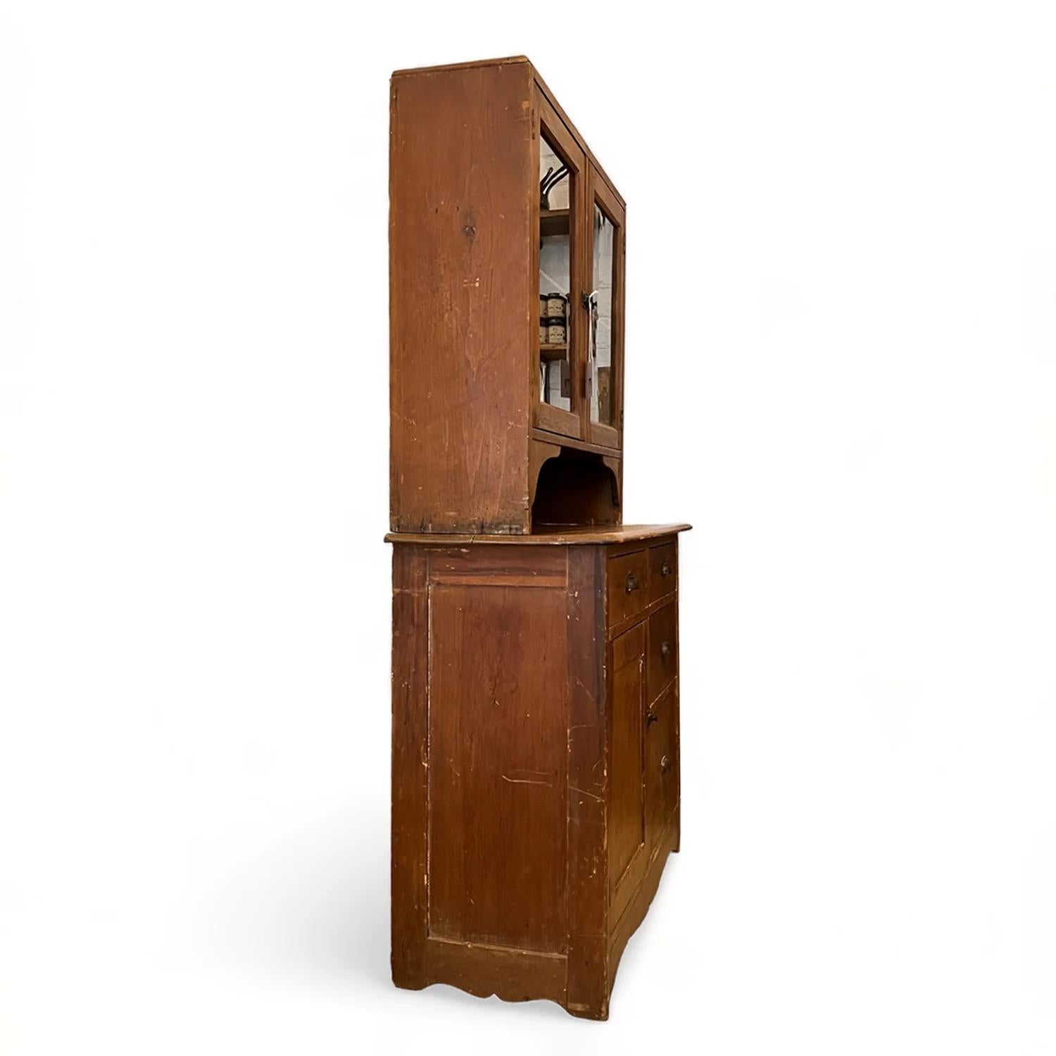 This timeless American farmhouse kitchen hutch cabinet, dating back to the early 20th century, boasts a rich patina and rustic charm. Its exquisite design includes an upper step-back hutch with two glass-paneled doors, providing a tasteful showcase