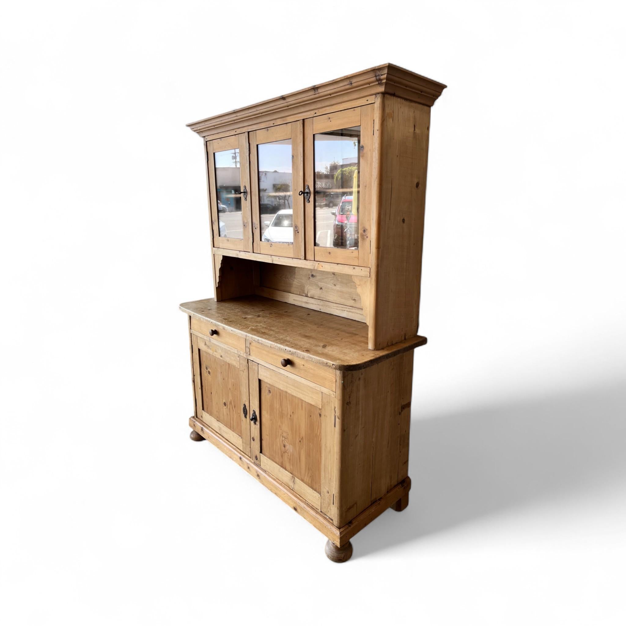 Este aparador vintage tiene unos encantadores armarios superiores con frentes de cristal, una amplia superficie de trabajo y almacenamiento inferior con dos cajones y puertas paneladas. Con su pátina rústica y su silueta clásica, ofrece