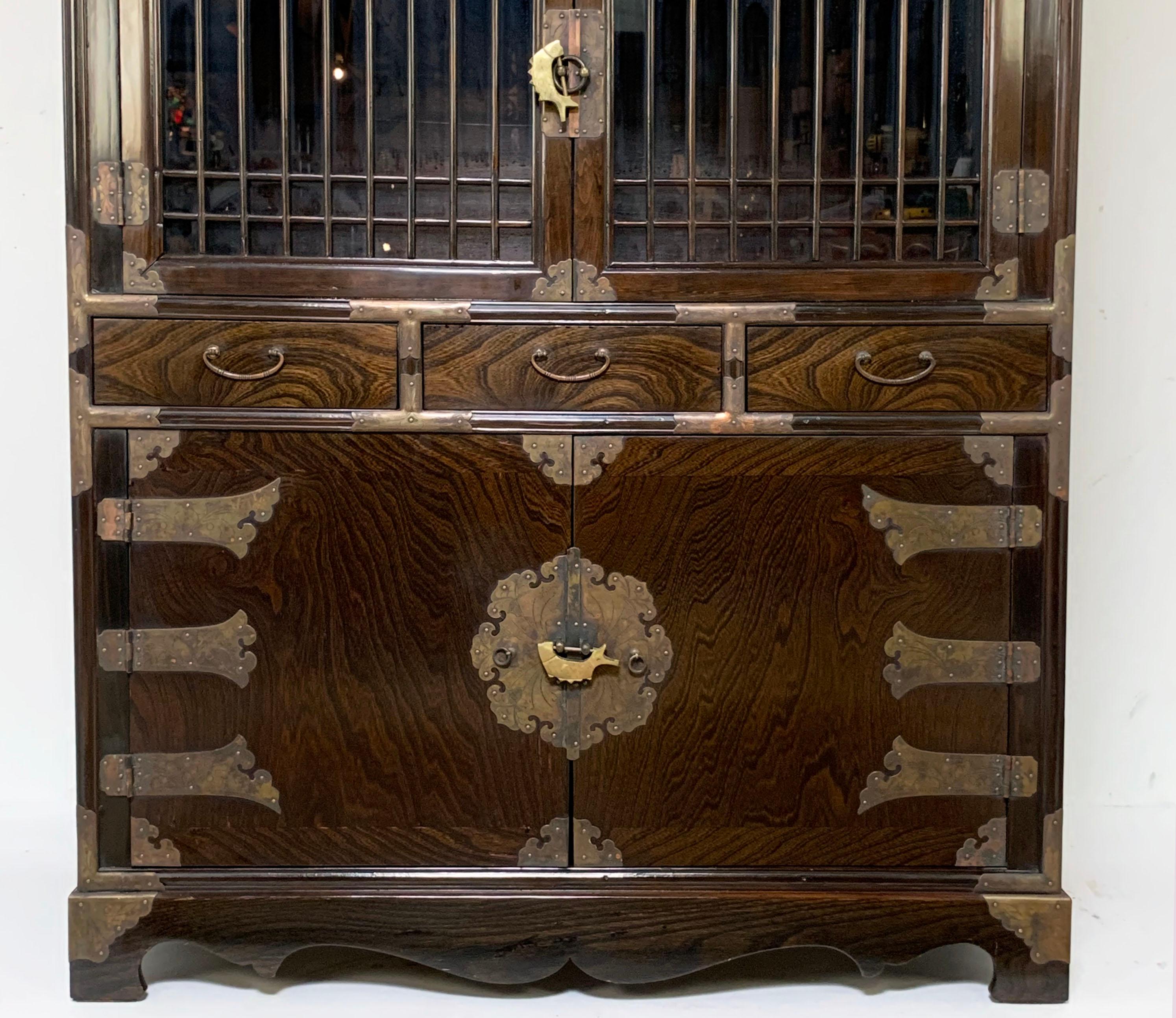 An unusual two-piece vintage Korean cabinet with glass display doors behind finely latticed mullions. Finely detailed brass work on this six foot tall display cabinet, comes with locks for all three sets of doors. 

The bottom cabinet measures
