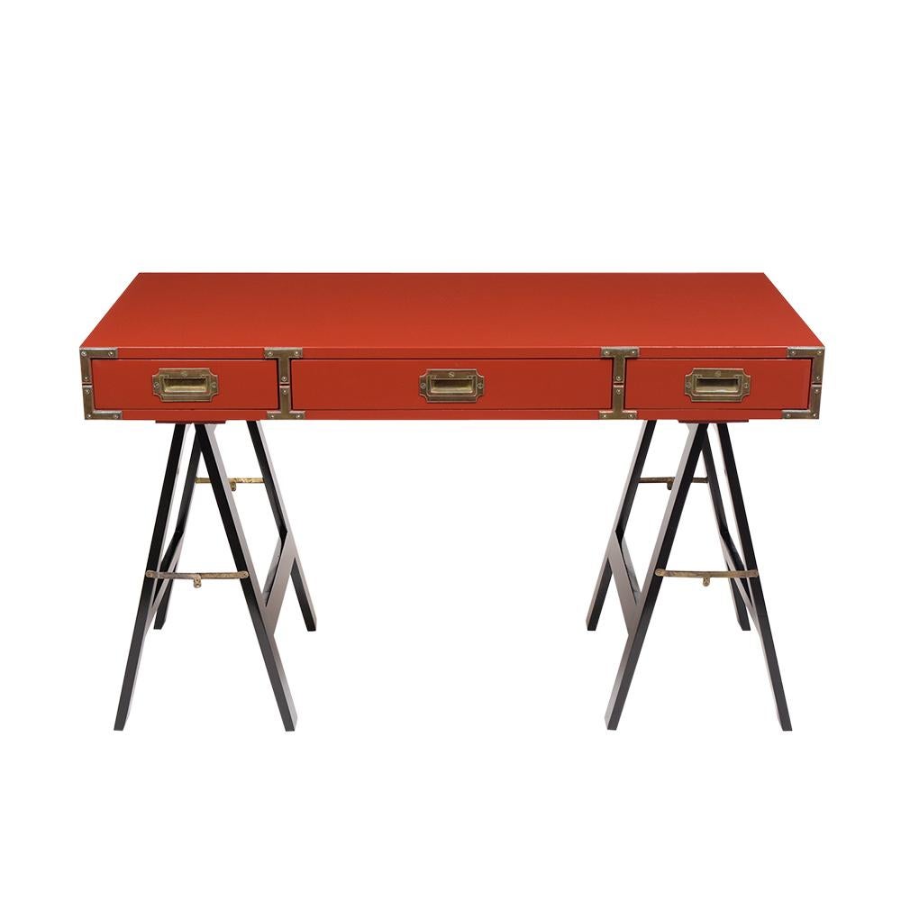 This midcentury Campaign desk has been restored and stained a red-orange color with a newly lacquered finish. The desktop has a semi-gloss finish, polished brass accents around the pullout / pull-out drawers, and rest on a pair of ebonized quad-pod