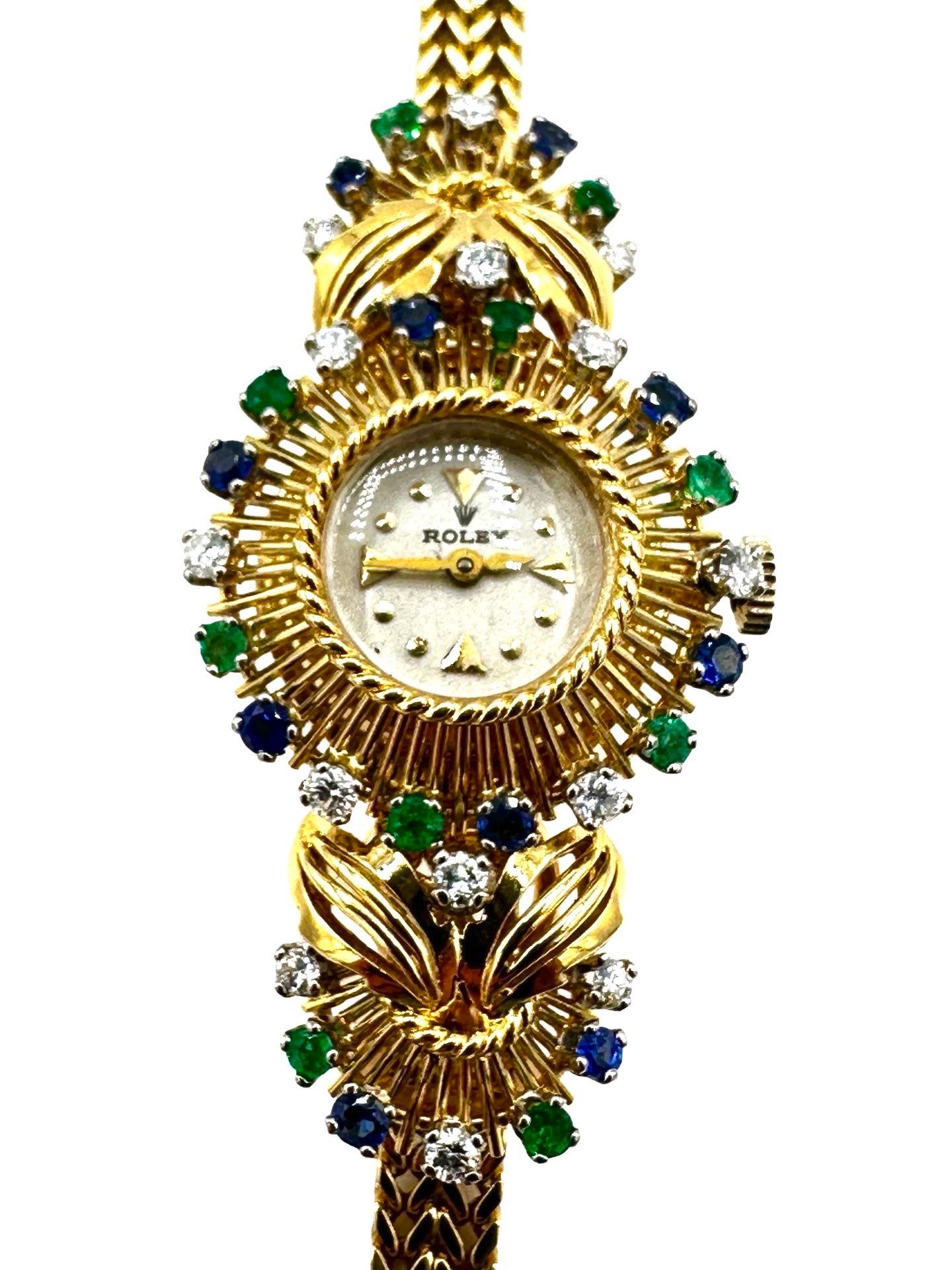 A rare vintage ladies Rolex 18 karat yellow gold watch with Diamond, Sapphire and Emerald accents.  The Diamonds, Sapphires and Emeralds are all round cut stones accenting a white center dial.  The two pieces next to the head of the watch, are