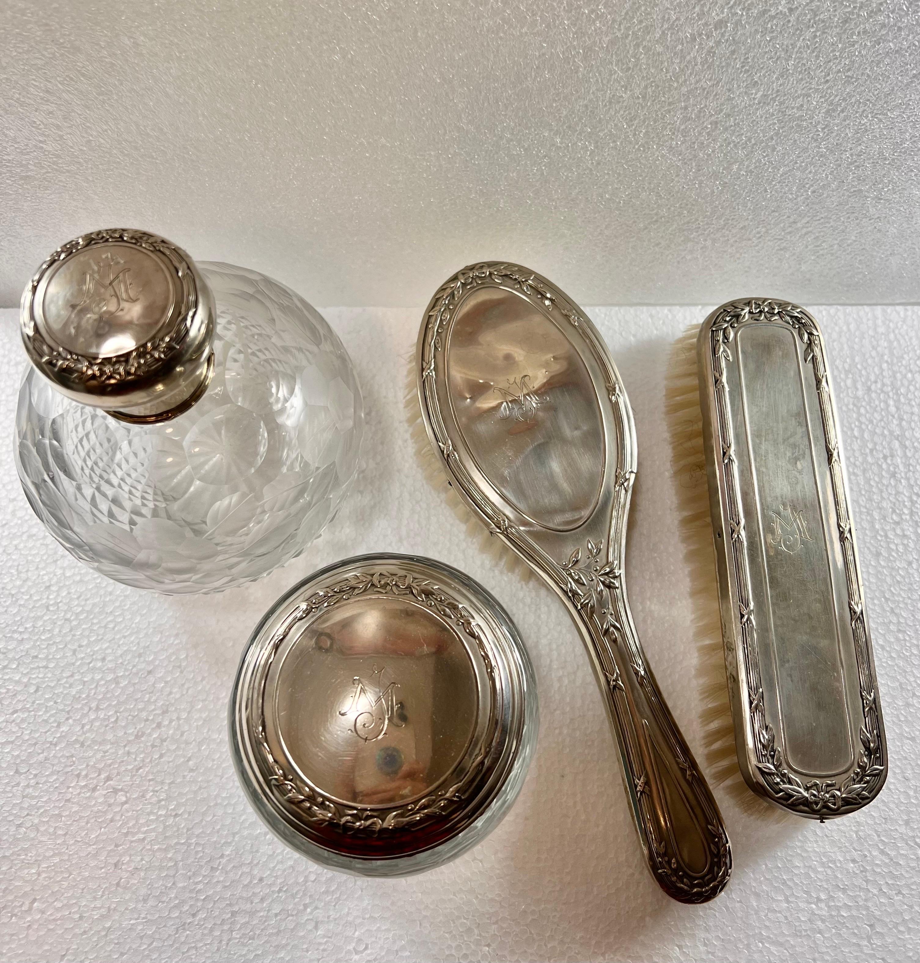 Four-piece vanity set in solid cut glass and silver. It includes a hand mirror with a beveled glass, one bottles, a clothes brush and a hair brush. The glass of jars and jars is solid and carved. All parts in silver present a stamped decoration and