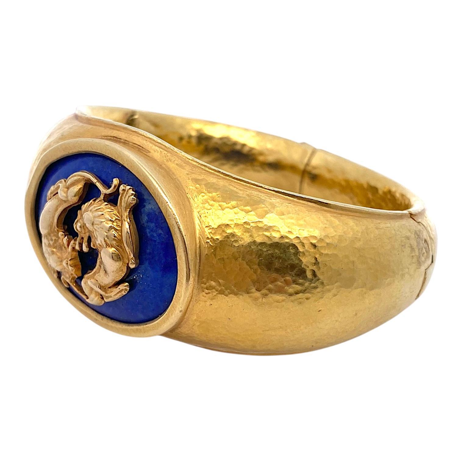 Vintage Ilias Lalaounis hammered 18K yellow gold double hinged bangle bracelet, featuring a central cabochon-cut lapis lazuli stone set within an oval bezel. Two intricately detailed gold lions encircle the stone, creating a dynamic focal