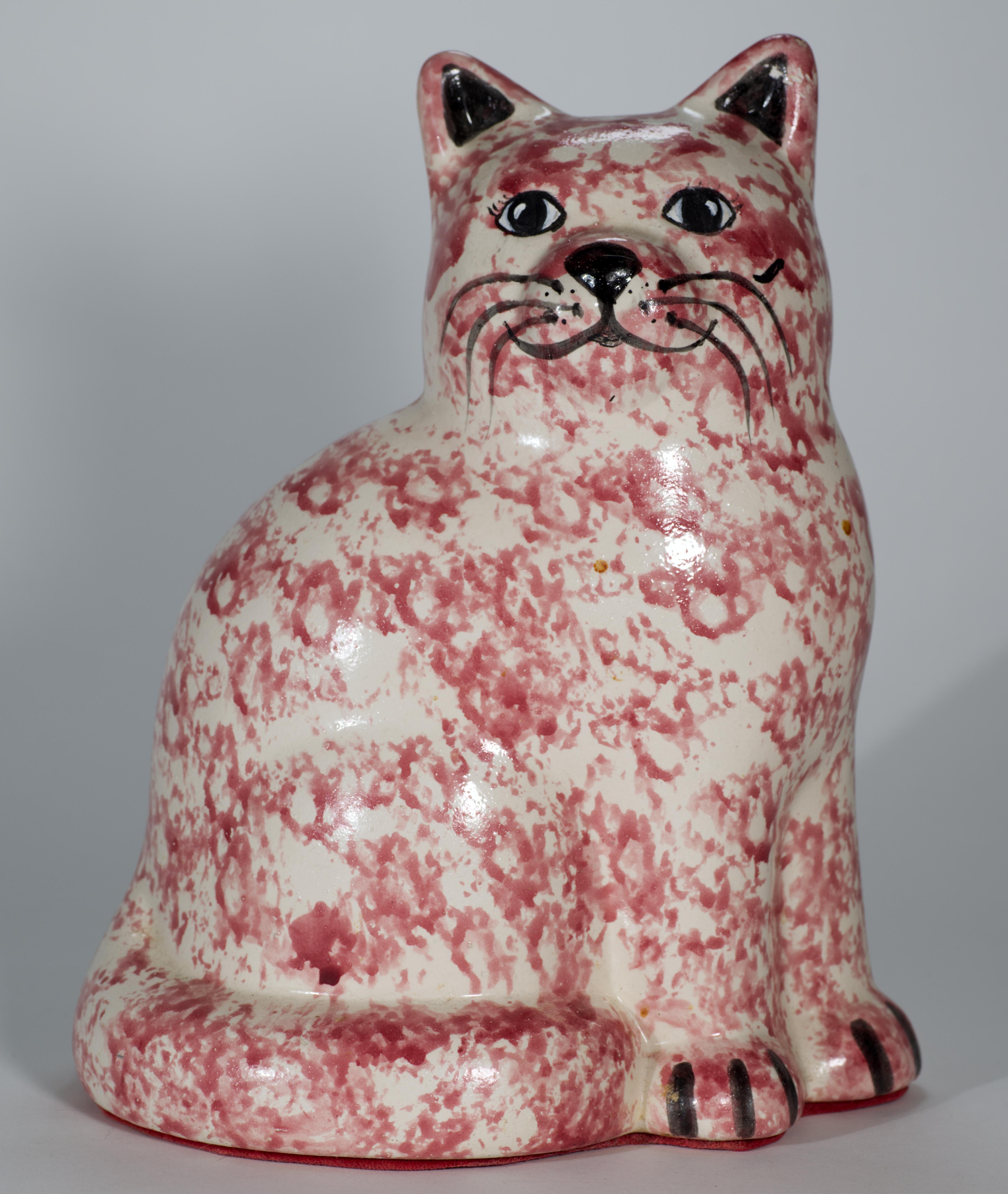 Vintage ceramic cat figurine features hand-painted pink sponge ware decor with black accents on face and paws. The cat depicted sitting with tail curled around the hind legs, slightly turned to the side and looking forward.

The figurine's bottom is