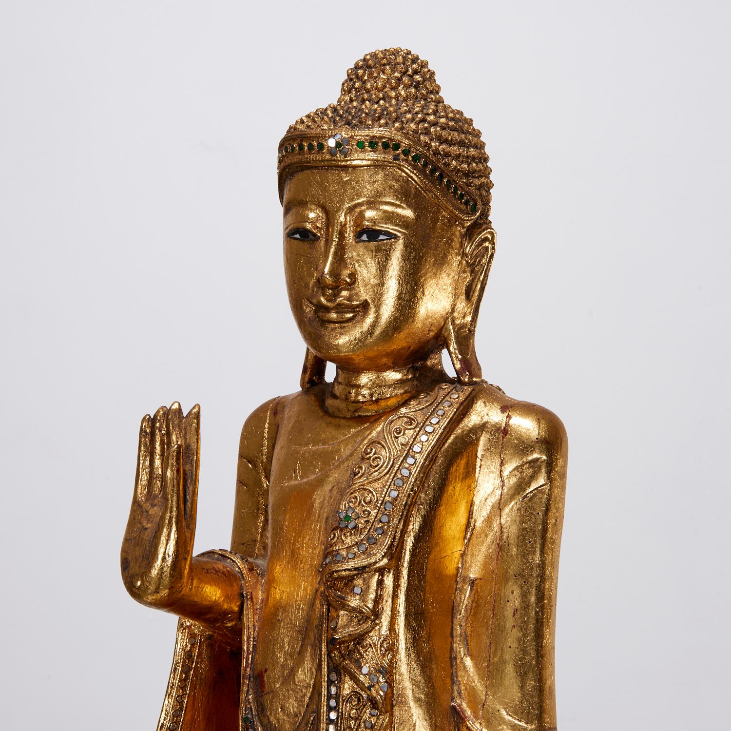 A lovely vintage 20th c., carved and giltwood standing Burmese Mandalay Buddha, with green and clear mirrored inlaid details, on wooden stand.

The Buddha is depicted standing with a palm held up facing outward signifying the act of teaching or