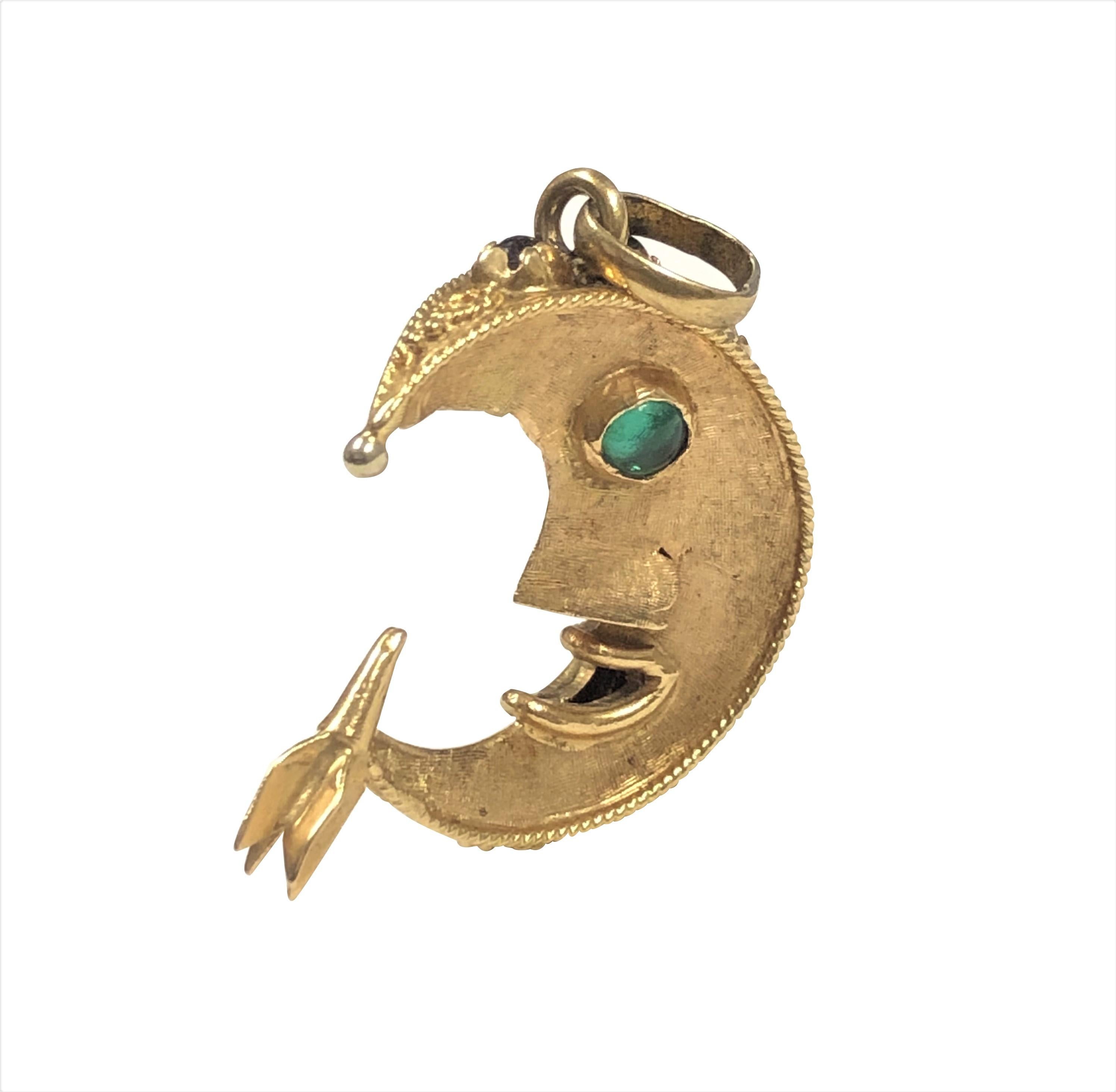 Circa 1970 18K yellow Gold Man in the Moon Charm , measuring 1 X 1 Inch X 3/8 inch thick and weighing 7.5 Grams, the 2 sided charm is lightly textured, nicely detailed featuring a Rocket Ship Landing at the tail of the Moon. Set with Red and Green