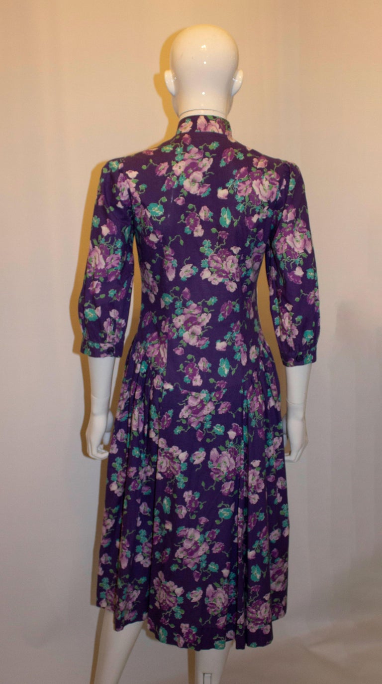 A pretty vintage floral cotton dress by Laura Ashley. The dress has a purple background with floral print, stand  up collar and button front.  It has elbow length sleaves with  cuff , a pocket on either side, and gathering at the waist.  UK size