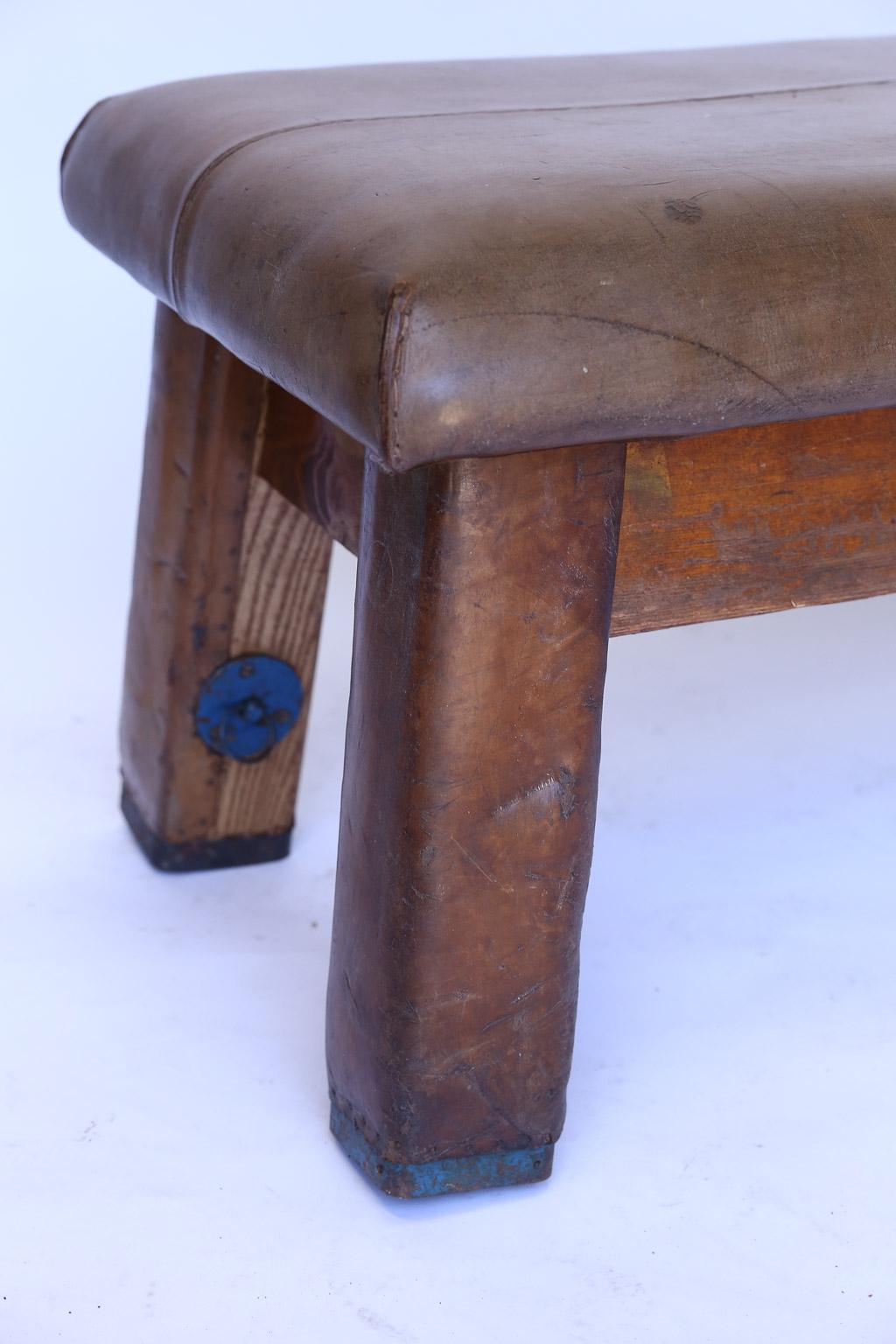 Found in France, this leather gym bench is most likely from the 1930s. It has a beautifully aged padded leather top on heavy wood legs with metal banded feet. Clever and perfect as a bench.