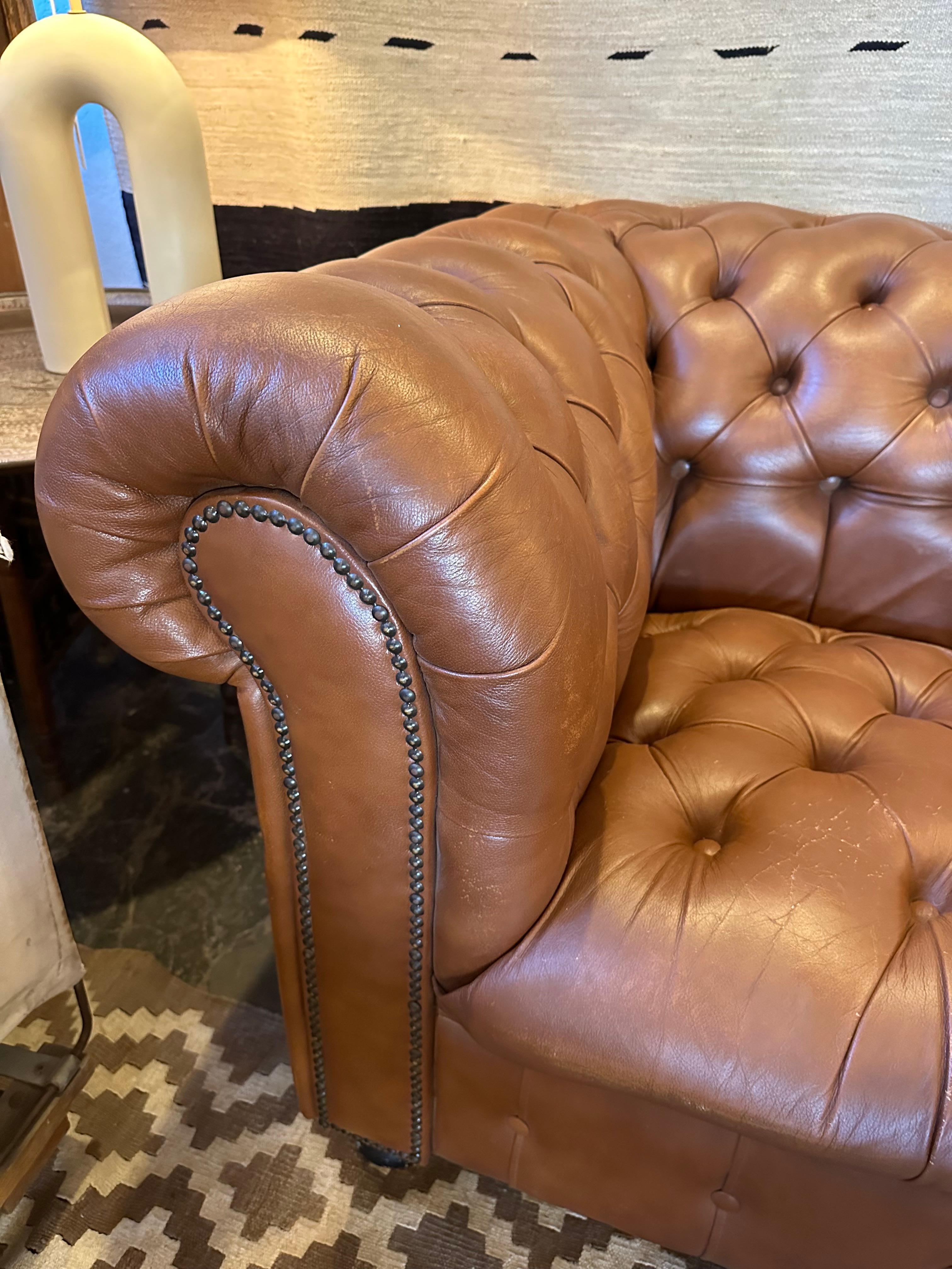 This vintage three seater Chesterfield stands as a timeless design staple, characterized by tufted leather upholstery and rolled arms. The leather, while softly aged, remains in good condition. Notably, it features the Empire Furniture Company