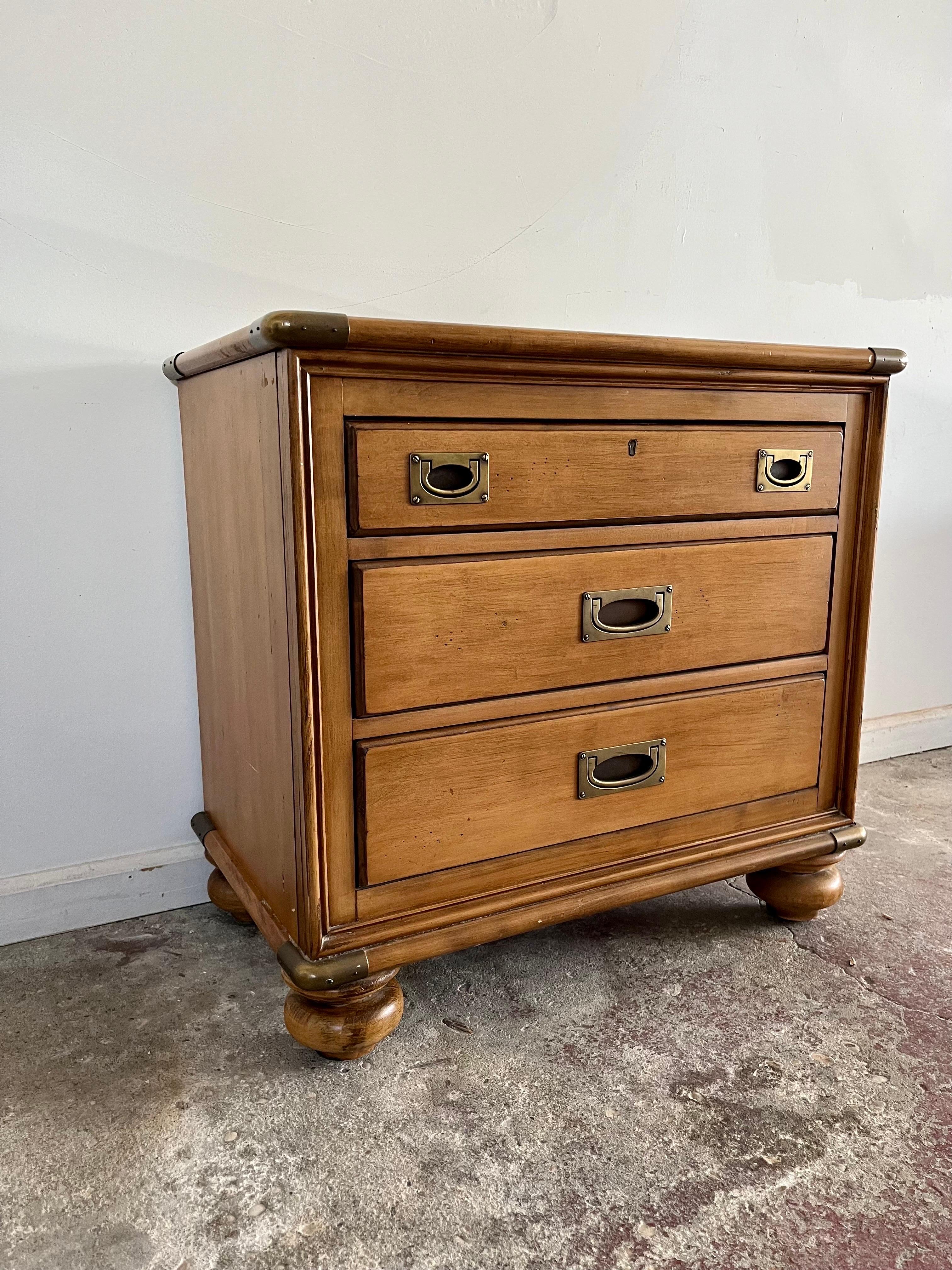 A light stained wood nightstand with brass accents by Lexington Furniture Nautica Home Collection. The chest features three drawers down the front each with brass handles, Brass capped corners and bun feet. The inside of the top right drawer reads