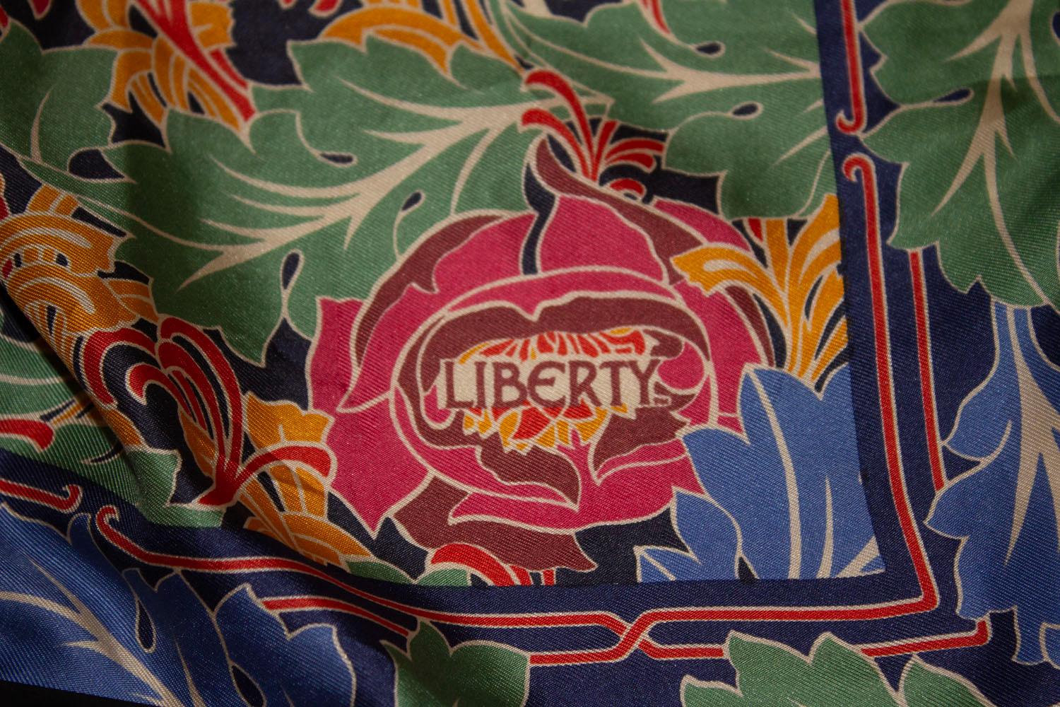 Un superbe foulard en soie vintage de Liberty of London. L'écharpe est bordée d'une bordure marine avec un motif de feuilles colorées. Les couleurs comprennent le rouge, le vert, le bleu et le rouge. Excellent état. Dimensions : 27'' x 27''