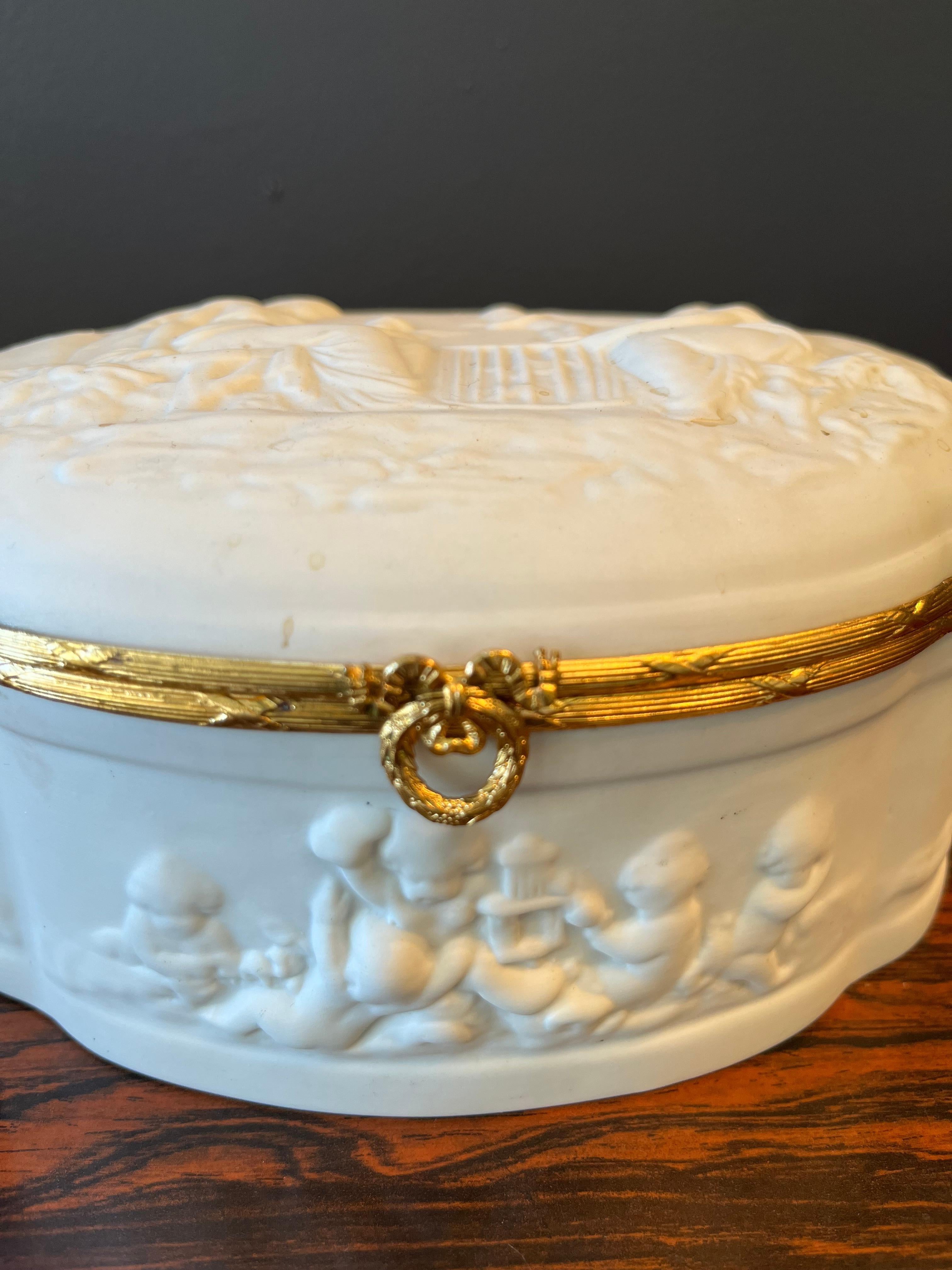 Ce magnifique cercueil vintage français de Limoges est fabriqué à la main en porcelaine blanche et présente un motif tridimensionnel d'anges dans un jardin.  Le dessin autour de la boîte de forme ovale présente des motifs plus tridimensionnels avec