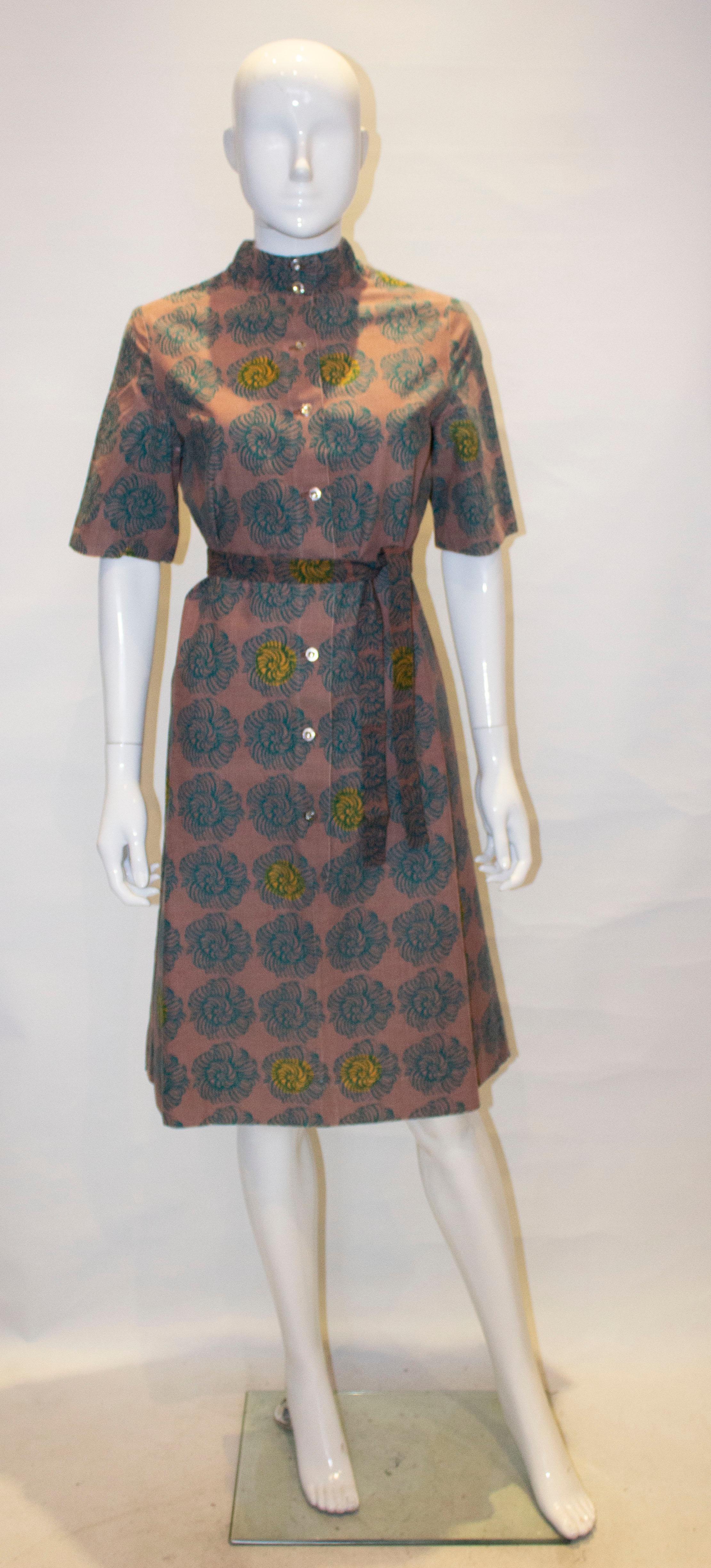 A great cotton dress by Maremeko of Finland from 1963. The dress has a light brown background, with a green and yellow print. It has short sleaves with a button front opening and self fabric tie belt. 