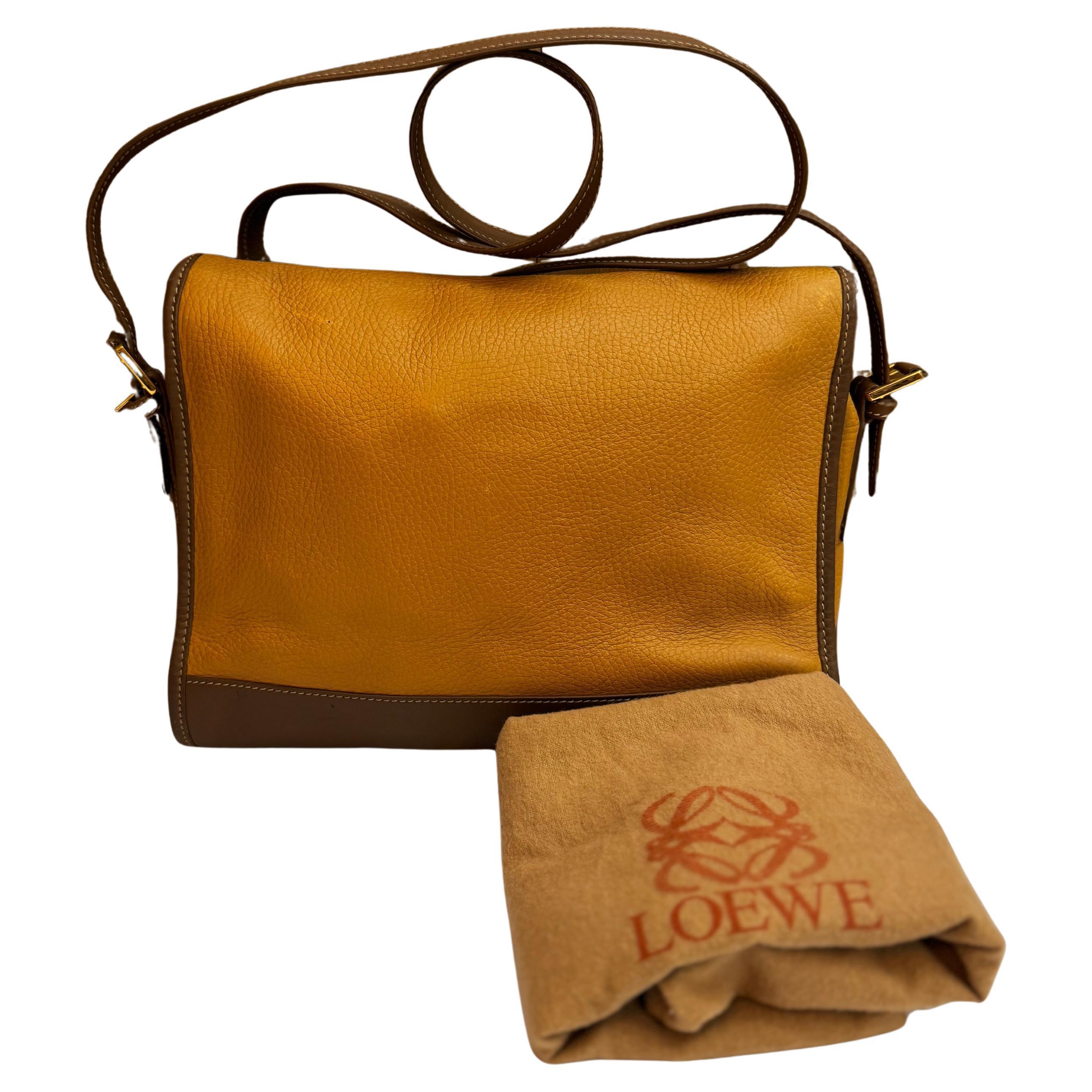 Ce sac photo vintage LOEWE Anagram est réalisé en cuir de veau à gros grain de couleur camel et garni de cuir lisse de couleur marron. Il est doté de ferrures dorées et d'une bandoulière du même cuir. La fermeture à bouton-pression magnétique du