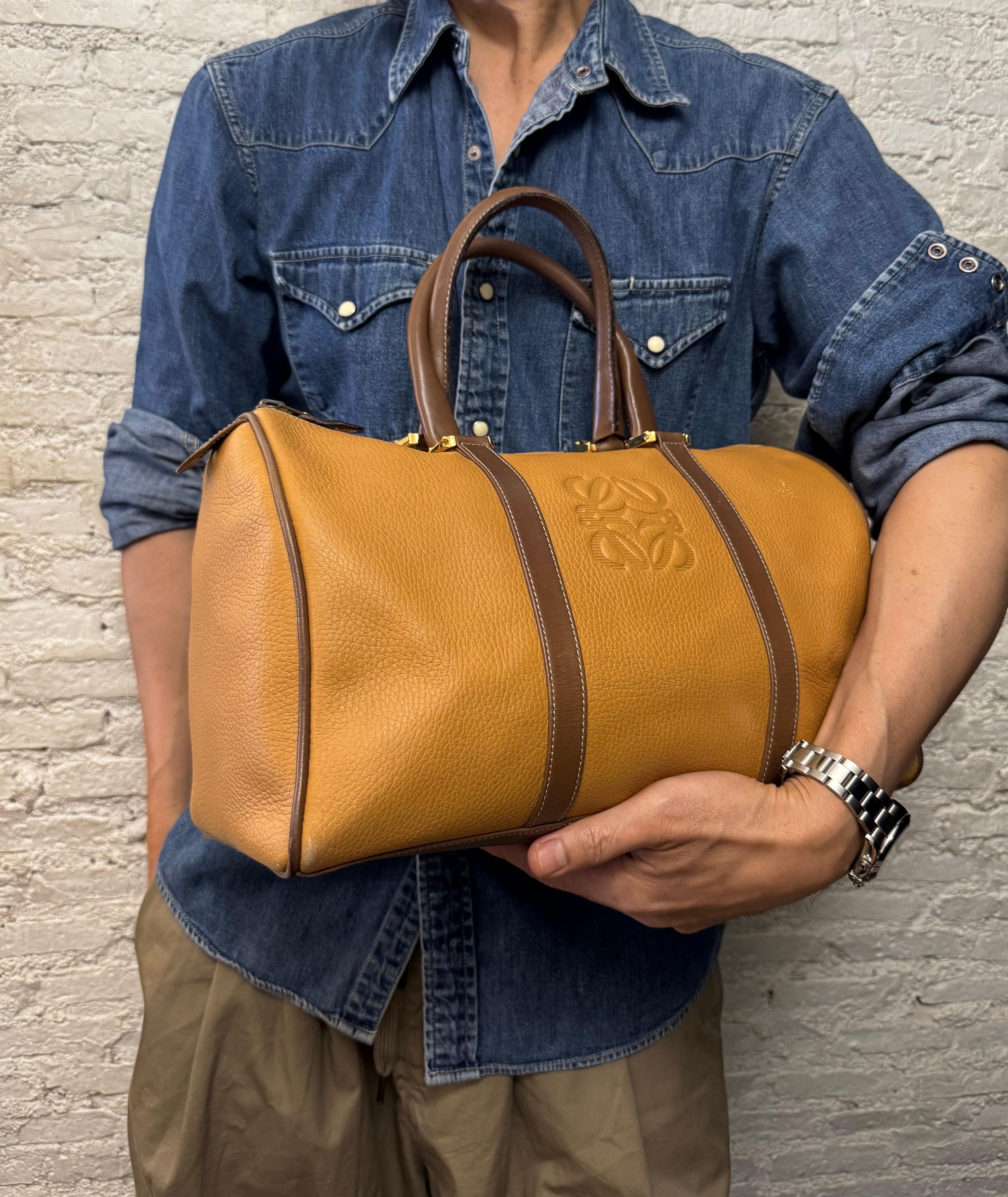 Diese LOEWE Anagram Boston Tasche im Vintage-Stil ist aus grob genarbtem Kalbsleder in Senfgelb gefertigt, das mit glattem Kalbsleder in Braun abgesetzt ist. Sie verfügt über goldfarbene Beschläge und gerollte Ledergriffe. Der Reißverschluss an der