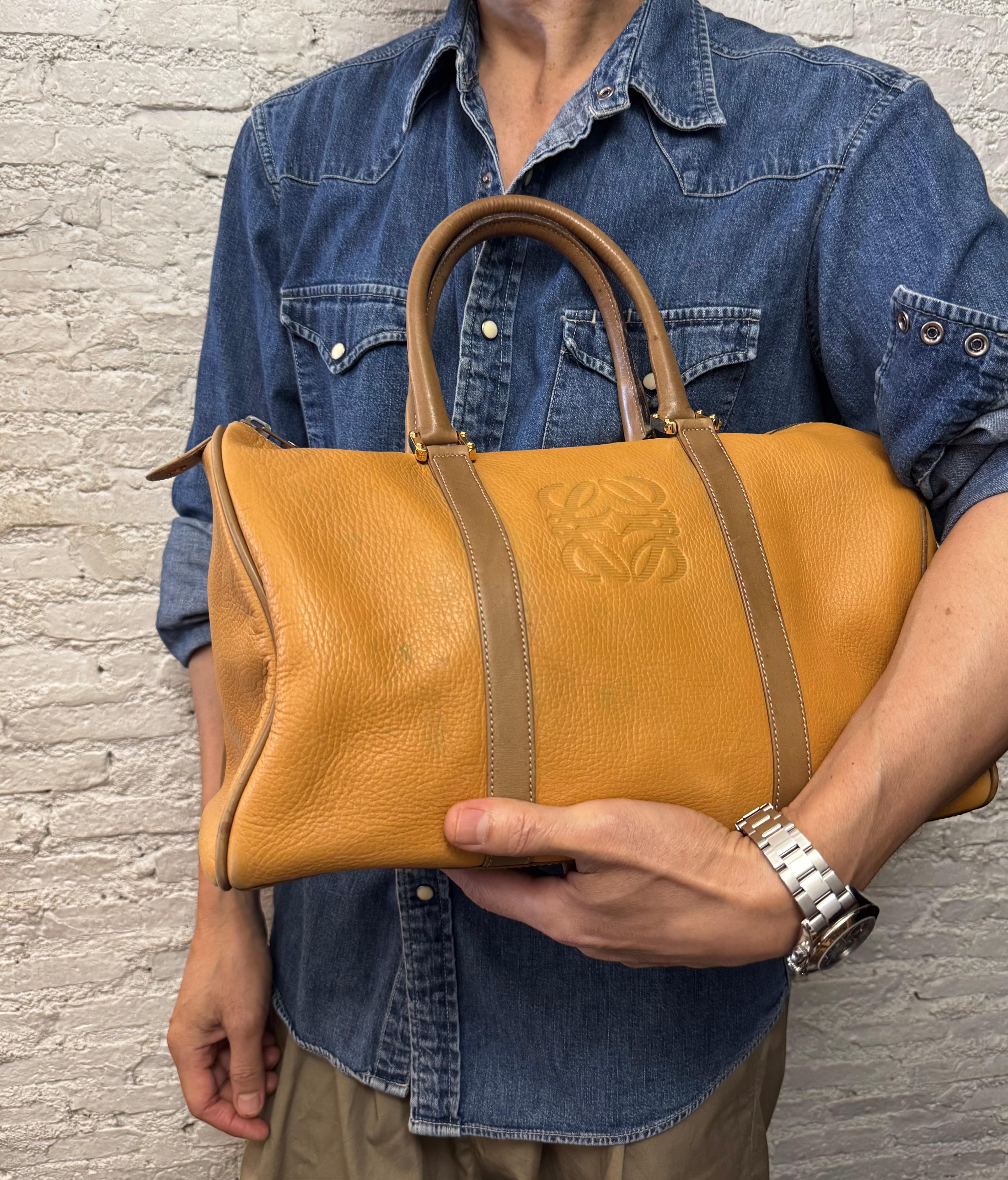 Diese LOEWE Anagram Boston Tasche im Vintage-Stil ist aus grob genarbtem Kalbsleder in Senfgelb gefertigt, das mit glattem Kalbsleder in Braun abgesetzt ist. Sie verfügt über goldfarbene Beschläge und gerollte Ledergriffe. Der Reißverschluss an der