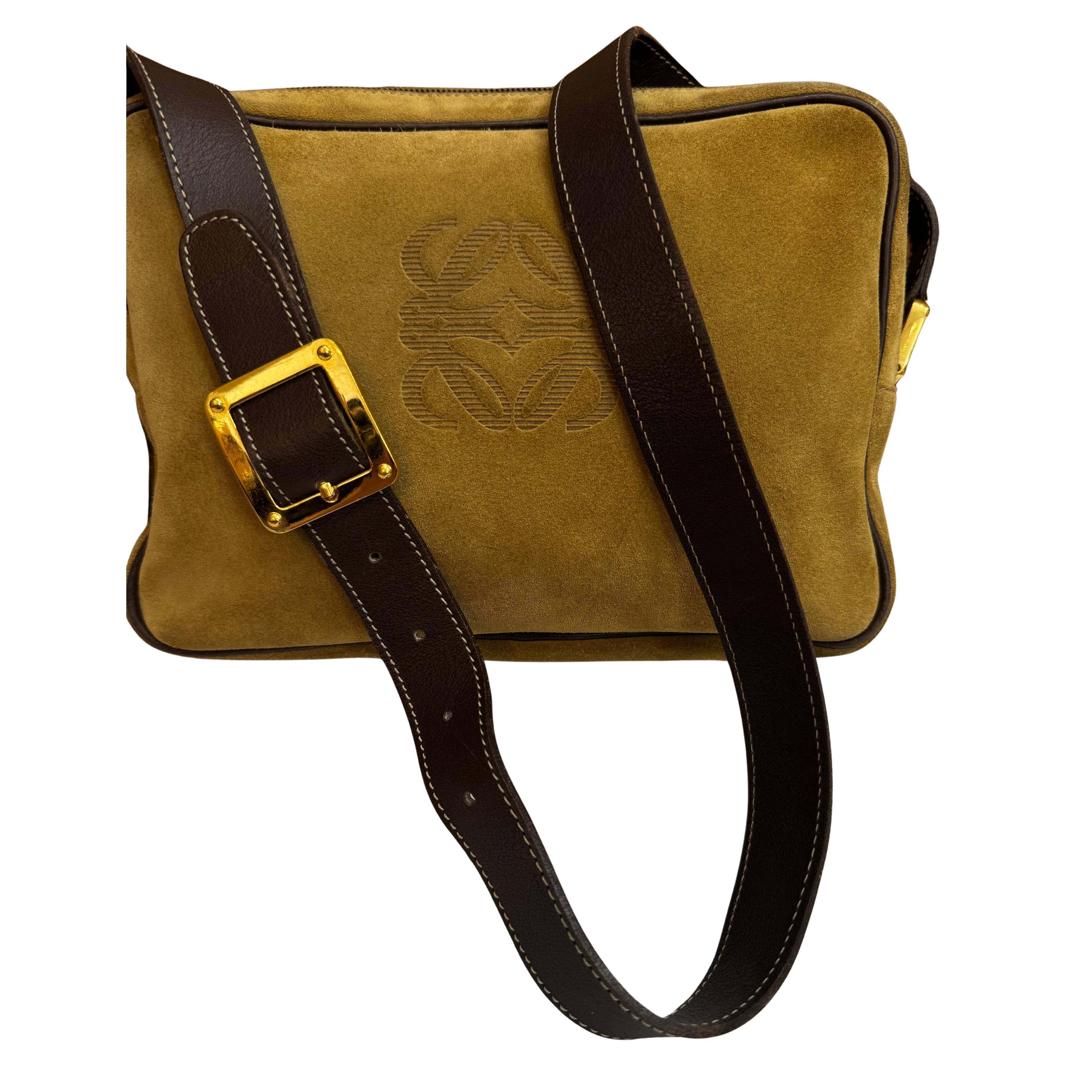 Este bolso vintage LOEWE Anagram para cámara está confeccionado en ante de becerro marrón con ribetes de piel lisa marrón oscuro, herrajes dorados y correa para el hombro de la misma piel. El cierre superior de cremallera se abre a dos