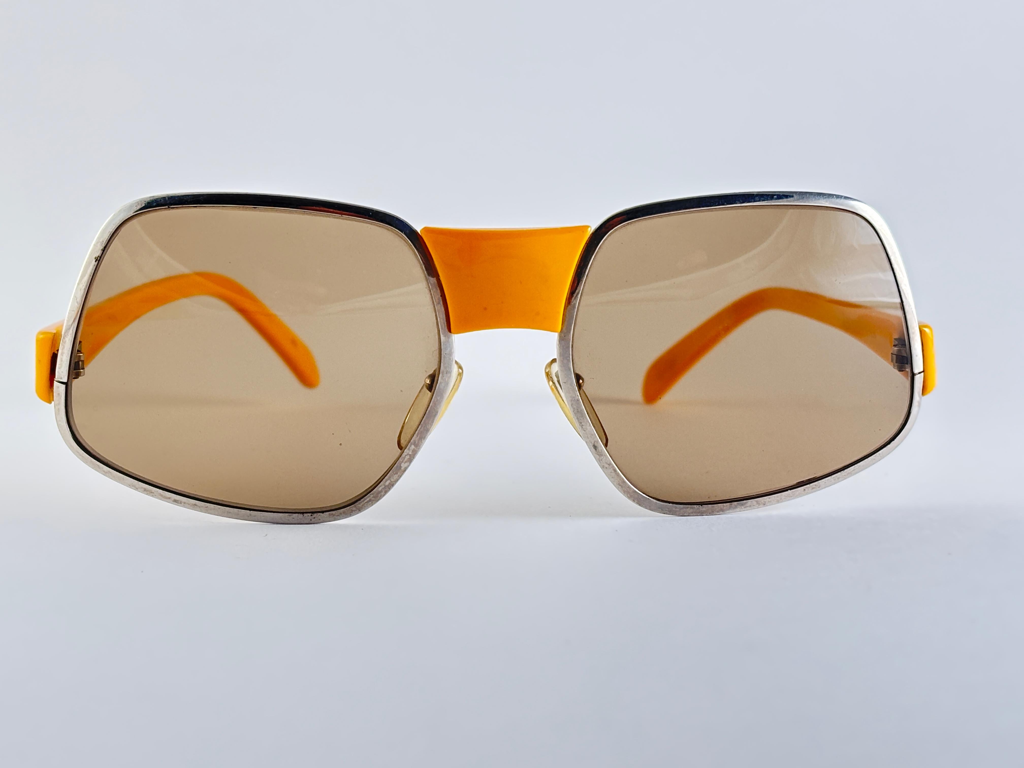 Übergroße Sonnenbrille in Metallic-Silber mit ockerfarbenem Nasensteg und Bügeln. 

Dieser Artikel hat kleinere Anzeichen von Verschleiß auf dem Rahmen durch die Lagerung.

Messungen 

Vorderseite : 13 Zentimeter 
Linse Höhe : 5.8 cm
Linse Breite :