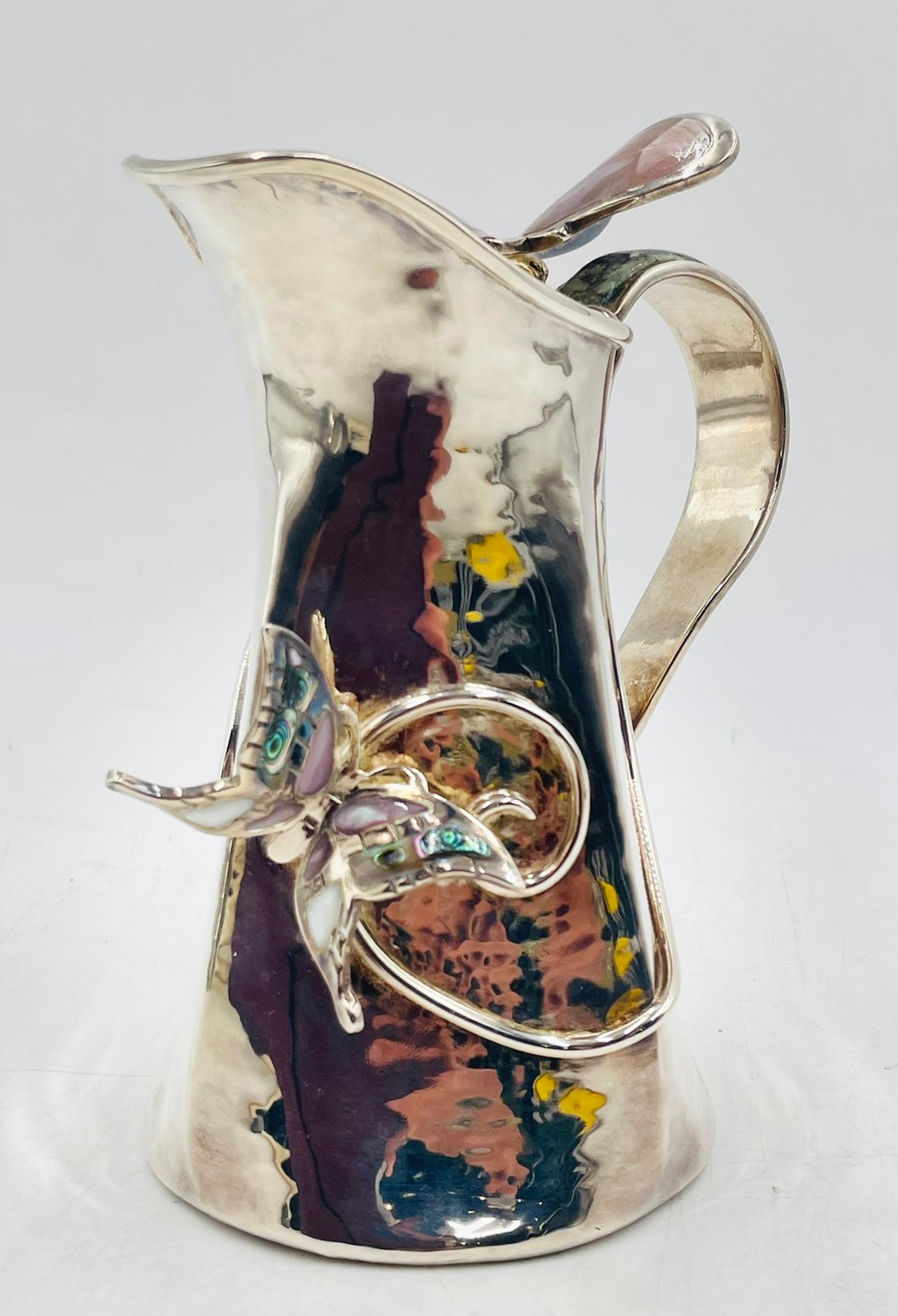 Excepcional jarra bañada en plata Los Castillo, de Taxco Mexico, con un caprichoso y artístico diseño de dos mariposas aplicadas: una con luminosas incrustaciones de nácar y la otra con vibrante concha de abulón. El mango escultural tiene