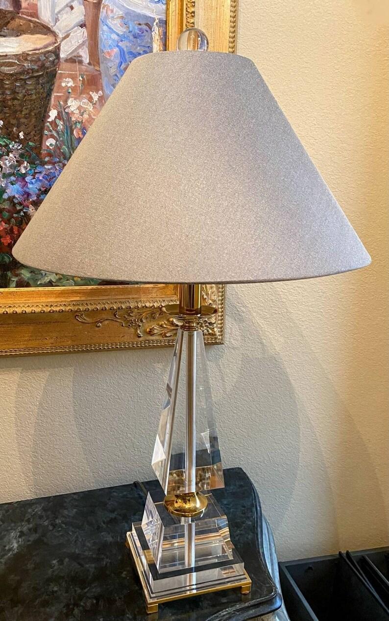 A fine geometric obelisk shaped lucite table lamp with gilt brass footed base, a grey-silver linen like shade, topped with a round lucite finial.

Dimensions:
30